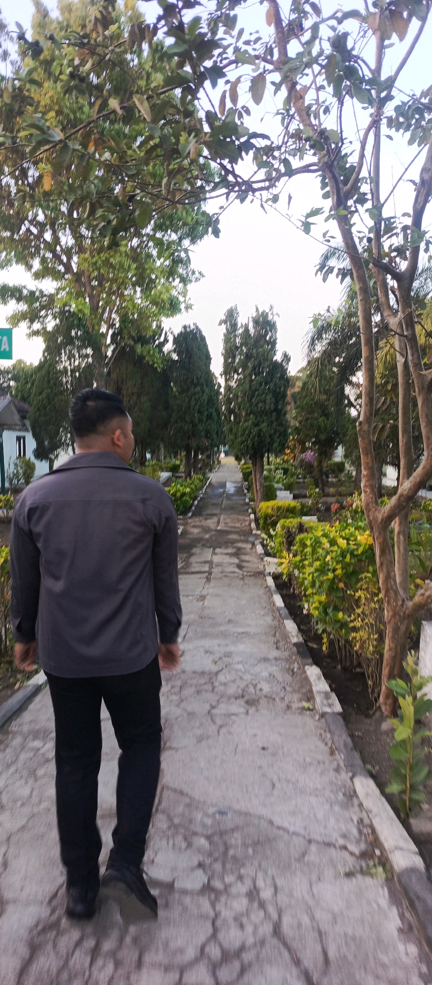 The image shows a man walking down a paved path in an outdoor setting. He is wearing a dark gray long-sleeved shirt and black pants. The path is bordered by lush green plants and trees. In the background, there are more trees, some of which appear to be cypress trees, and a building with a white facade. The sky is visible at the top of the image and appears to be overcast or nearing sunset. The scene likely depicts a park or garden area within the city of Batu, Indonesia, known for its natural beauty and tourist attractions.