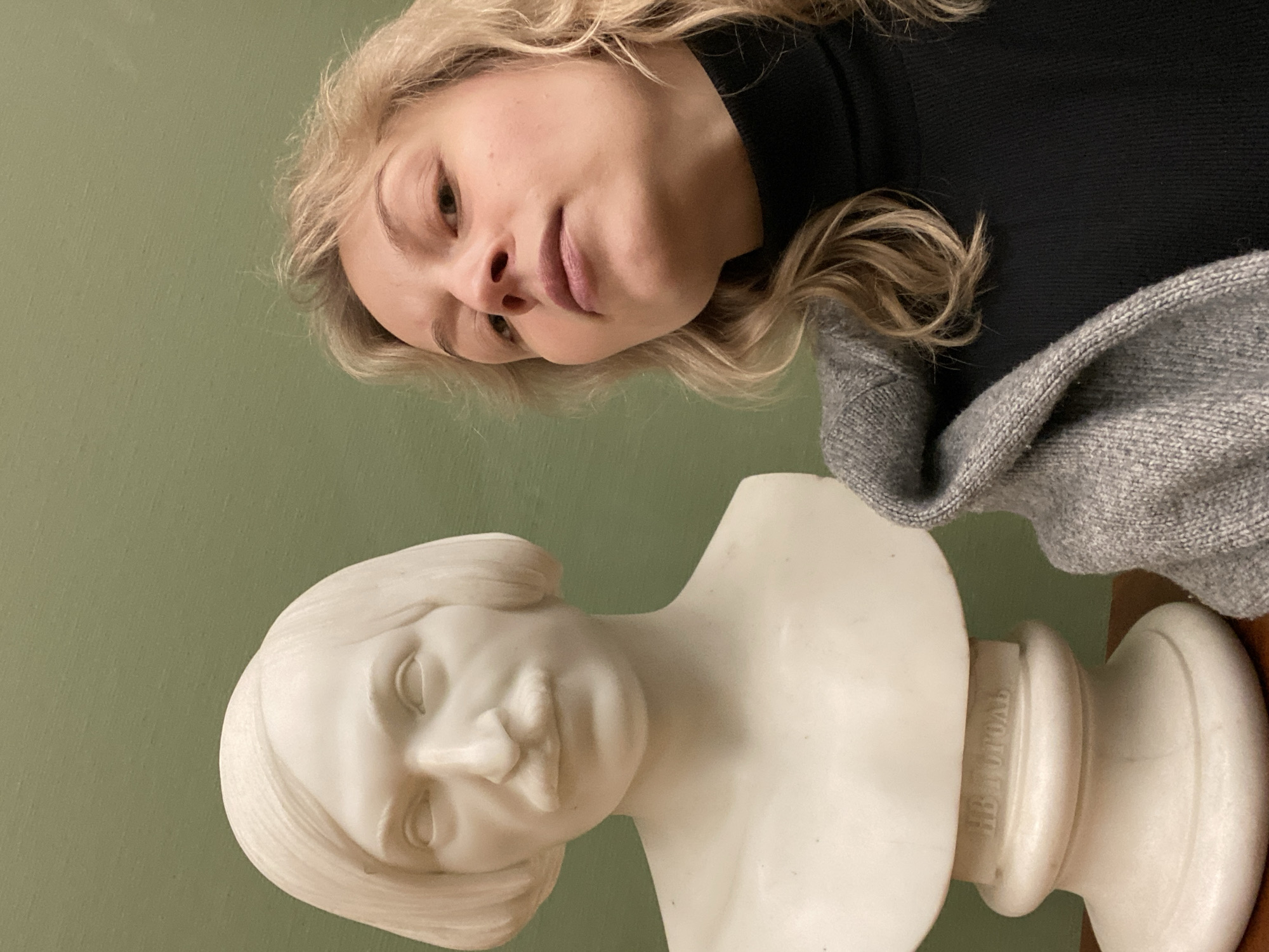 A portrait-style photograph is taken indoors, focusing on a fair-skinned woman with blonde, wavy hair and a bust of a marble-like statue. The woman is wearing a black turtleneck sweater with a gray knit sweater draped over her shoulders. Her gaze is directed downwards towards the statue. The statue appears to be of a person with short hair, resting on a cylindrical base with what appears to be lettering in Cyrillic script. The background is a solid green. Based on the Cyrillic script on the base of the statue, the location is possibly the city of Moscow, Russia.