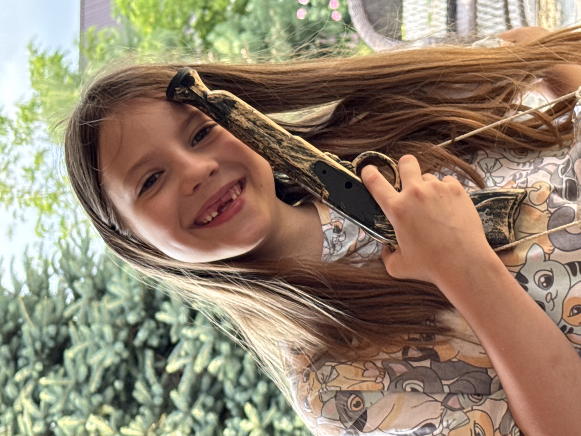 In this close-up, slightly tilted, outdoor photograph, a young girl with long brown hair is holding a toy gun. She is smiling broadly at the camera, revealing a gap where a tooth is missing. Her hair is brushed back from her face, with some strands falling forward. She is wearing a light-colored shirt with a repeating pattern of cartoon cats. The toy gun has a brown and black camouflage design.
The background is blurred, but appears to be a backyard setting with green foliage, possibly a tree or bushes, and hints of a building. The lighting suggests it is daytime, likely a sunny day. The girl's expression is joyful and playful. There is no visible text in the image. The location context is Zassovskaya, Russia.