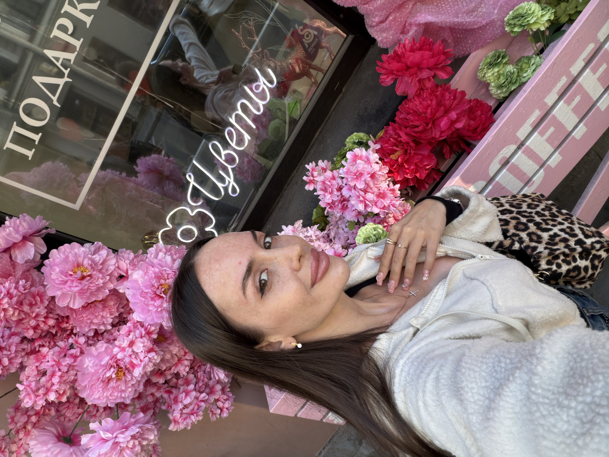 A young woman with dark hair and freckles is taking a selfie in front of a flower shop in Saint Petersburg, Russia. She is wearing a cream-colored fleece jacket and a necklace with a cross pendant. The flower shop window displays pink and red flowers, a neon sign that reads "Цветы" (Flowers), and a golden deer figurine. The shop is decorated with pink accents, including a bench with the word "OFFER" painted on it.