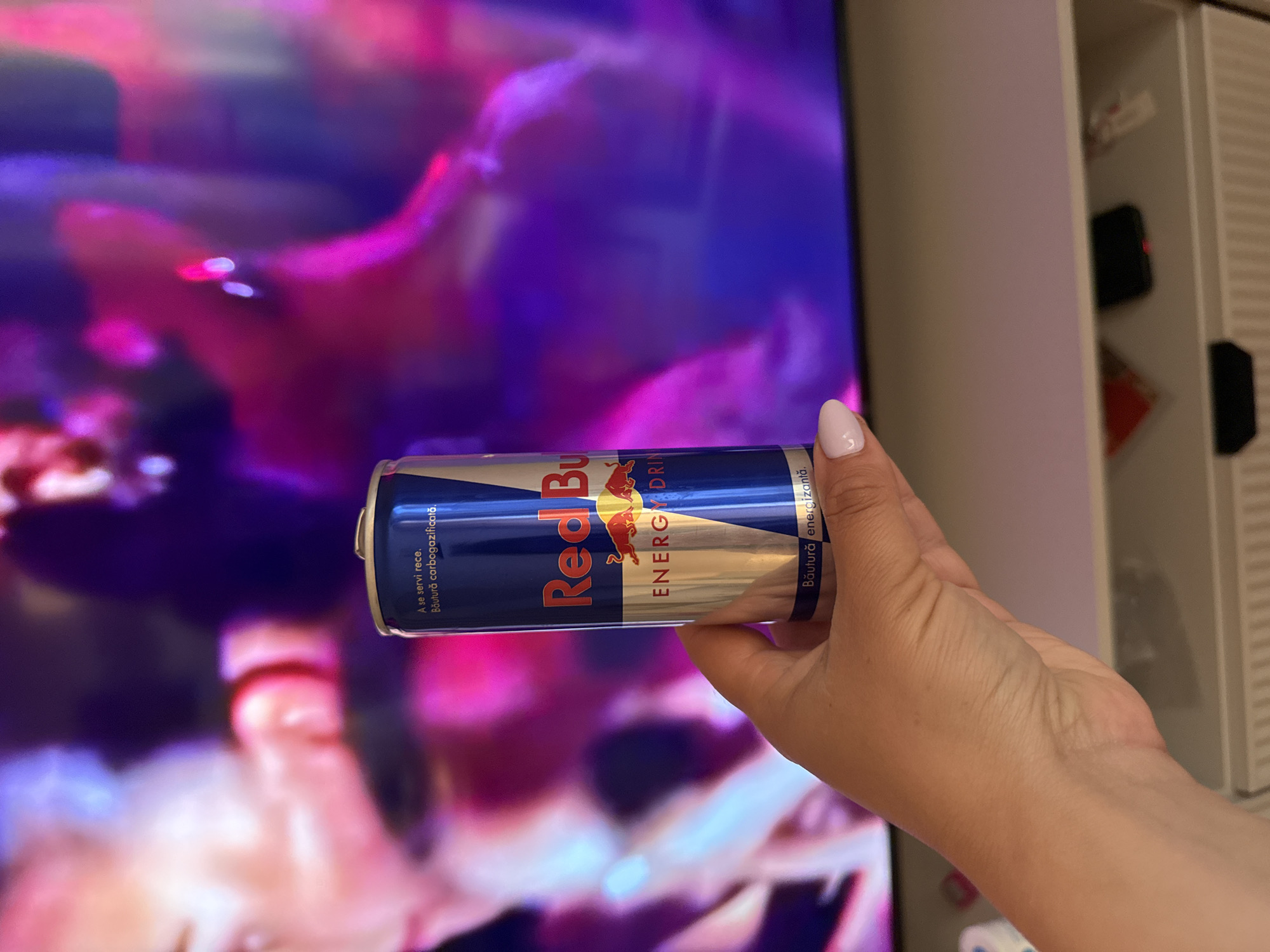 A hand is holding a can of Red Bull energy drink in front of a blurry television screen. The background includes a white cabinet with black objects inside. The text on the can is in Romanian.