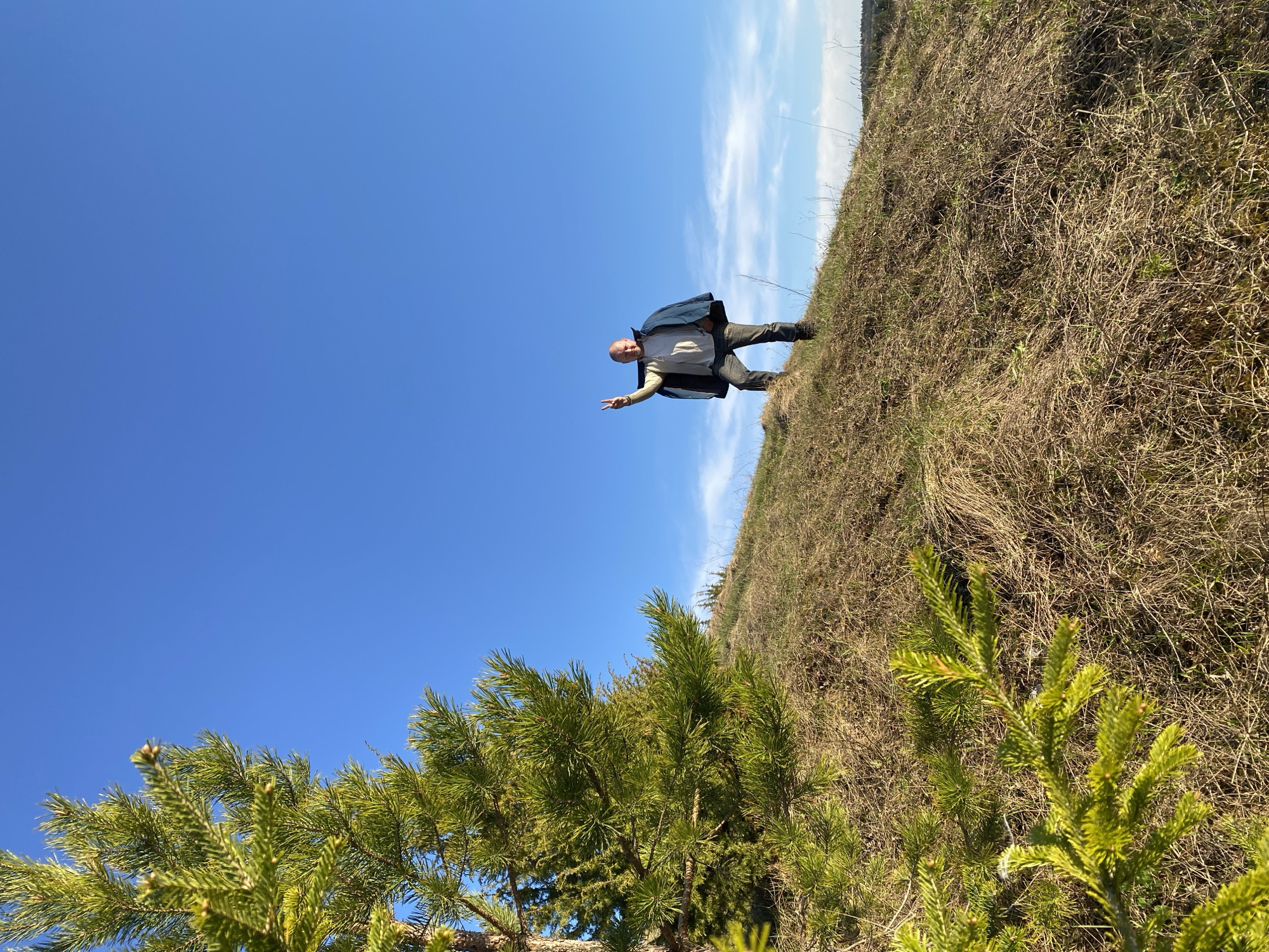 The photograph shows a man standing on a grassy hill in Ольховка, Russia. The man is bald, wearing a jacket, pants, and shoes. He is extending his hand in a "peace" sign. The hill is covered in brown grass and some green vegetation. The sky is blue with wispy clouds. A pine tree is visible in the lower left corner.