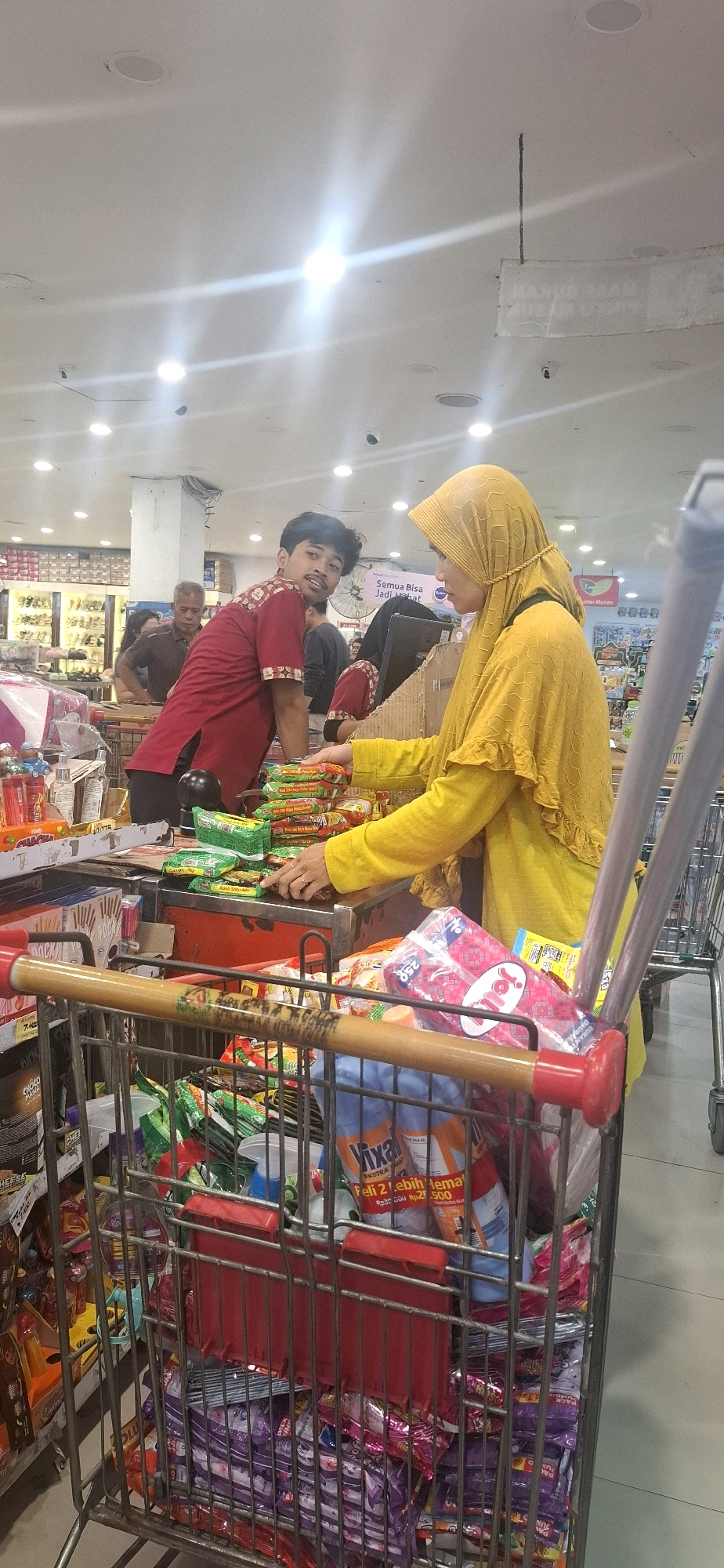 The image depicts the interior of a supermarket, likely in Kartasura, Indonesia, based on the text on some of the products. Two individuals are visible near a checkout counter. A man wearing a maroon shirt with a patterned design is leaning over the counter, looking towards the viewer. A woman wearing a bright yellow hijab and a yellow shirt is standing and appears to be interacting with the man. She is holding a stack of small packets, possibly instant noodles, and is reaching out with her left hand. In the foreground, a shopping cart is filled with various grocery items, including packaged snacks, bottles, and what appear to be cleaning supplies. The store environment features bright overhead lighting, shelves stocked with merchandise in the background, and tiled flooring. The overall scene suggests a typical shopping interaction at a retail establishment.