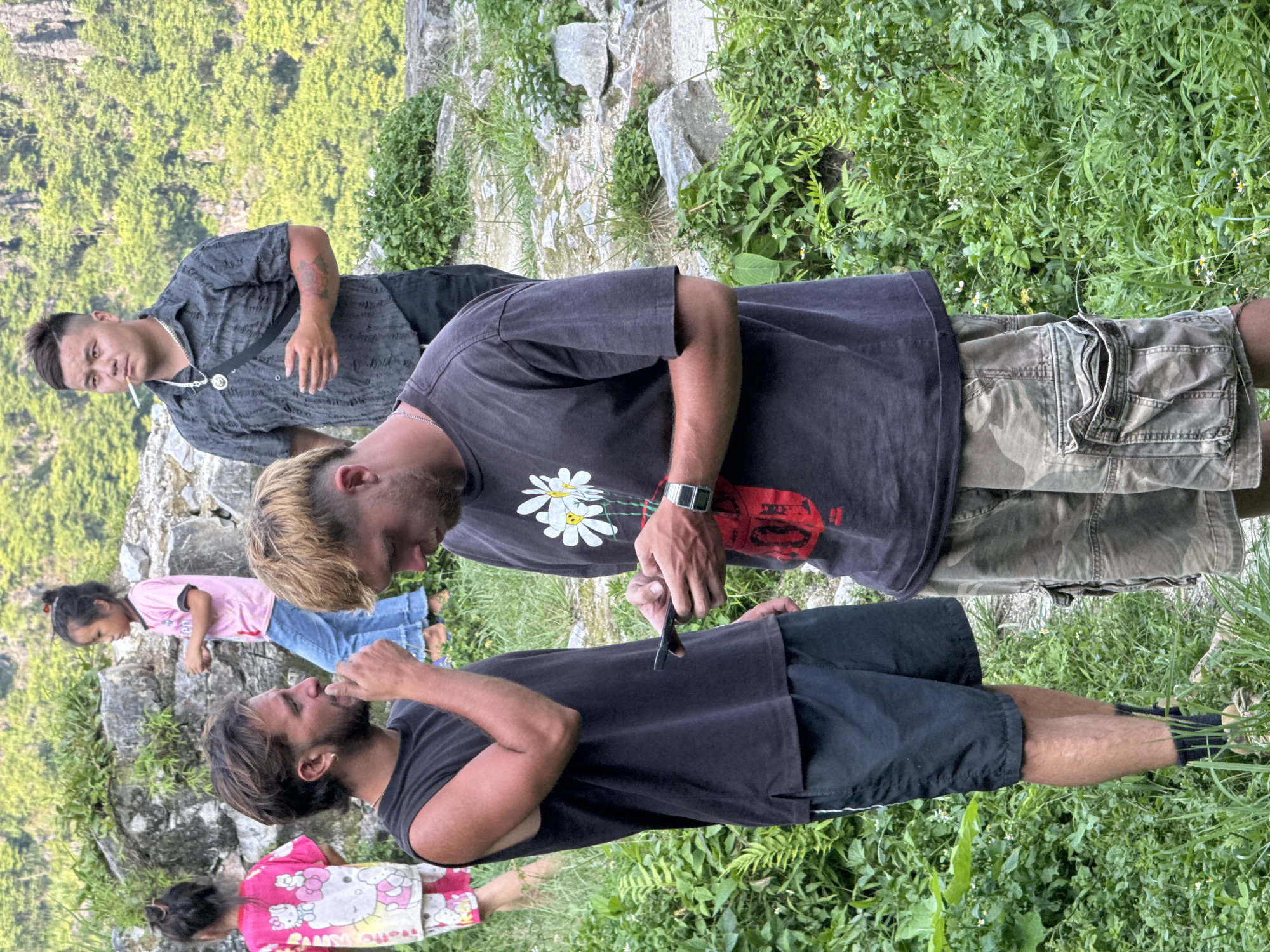 In Đường Thượng Commune, Vietnam, a group of individuals is gathered in a natural setting. A man in a gray patterned shirt stands near the edge, holding a cigarette. Closer, another man with blond hair looks down at a smartphone or similar device. Next to him, a third man with dark hair raises his hand to his mouth. Two young girls are present; one wears a pink shirt and jeans, while the other is dressed in a pink Hello Kitty shirt. The environment features greenery, rocks, and vegetation. The setting appears to be a hillside with a steep, forested slope in the background.