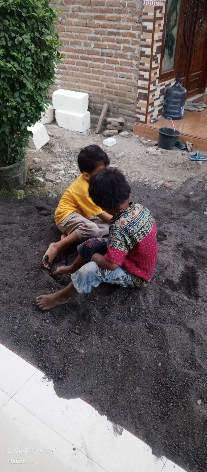 In the city of Lebanisuko, Indonesia, two young boys are observed sitting on a pile of dark soil. The boy on the left is wearing a yellow t-shirt and light-colored pants, with his bare feet stained with the soil. The boy on the right is wearing a colorful, patterned shirt and denim shorts, with his bare feet also in the soil. They appear to be engaged in an activity together, likely playing with the dirt. The background includes a brick wall, a large green bush in a pot, and a doorway with a water cooler and buckets nearby. The ground is a mix of soil and gravel, with some white blocks and debris scattered around. A tiled pathway is visible in the foreground. The timestamp "2026/08/19 17:24" and the name "Hafizh" are superimposed on the image.