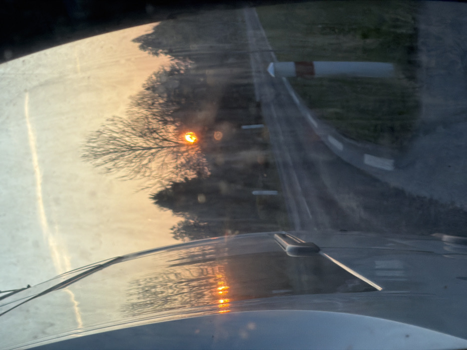 The image, captured from within a vehicle in Le Saulcy, France, presents a rural road scene during either sunrise or sunset. No individuals are present. The left foreground is occupied by the light-colored side body panel of the vehicle, featuring a dark-colored door handle and reflecting multiple instances of the bright orange sun and surrounding bare trees. The view through the vehicle's streaky window reveals a paved road, marked with a white dashed line, curving sharply to the right. A grassy embankment lines the roadside, where several white bollards with orange bands are positioned. In the background, a dense line of mostly bare deciduous trees and some evergreen foliage is silhouetted against a sky displaying gradients of orange and yellow near the horizon, transitioning to light blue overhead. The sun, an intense orange-yellow disc, is low on the horizon, partially obscured by tree branches, and exhibits lens flare effects. A faint contrail is visible in the upper sky.