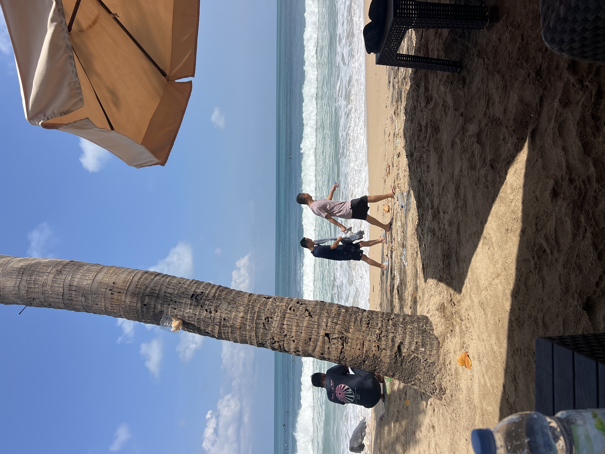 The image depicts a sunny day at Kuta Beach, Indonesia. A large coconut palm tree stands prominently in the foreground, its rough bark textured by time. To the left of the tree, a person is crouched on the sand, facing away from the camera, wearing a dark t-shirt and shorts. Further down the beach, two men are walking towards the right. The man in the middle wears a dark blue t-shirt and shorts, carrying a gray bag, while the man to his right has on a light pink t-shirt and black shorts, holding a clear plastic bottle.
The ocean stretches out in the background, with gentle waves rolling towards the shore. The water is a vibrant turquoise, contrasting with the white foam of the breaking waves. Several surfers can be seen in the distance, enjoying the ocean. Above the beach, a large beige umbrella provides shade, partially obscuring the bright blue sky dotted with a few white clouds. The sand on the beach is a warm, golden hue, with scattered debris suggesting recent activity. To the right of the frame, a dark wicker-style table is visible, holding a black object, likely a bag or cushion. In the lower-left corner, a partially visible plastic water bottle sits on a dark surface, suggesting a resting area or cafe. The overall scene captures a relaxed atmosphere at a popular tourist destination.