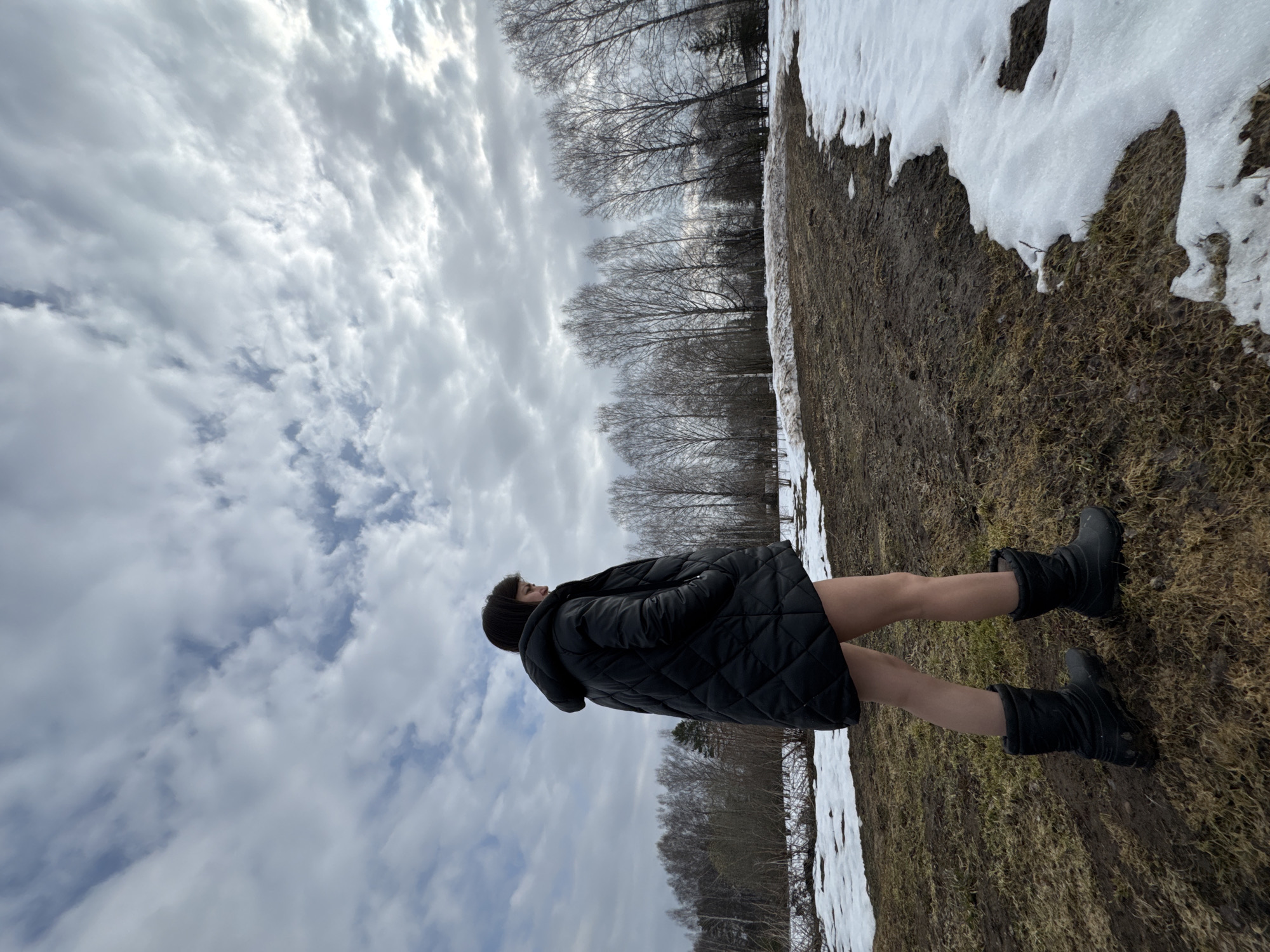 The image captures a woman standing in a field in Вассята, Russia, on a day with patchy snow. The ground is brown with grass, indicating the beginning of spring. The woman is wearing a black quilted jacket and black boots. The background shows bare trees against a cloudy sky. The photo is taken at a low angle, with the camera tilted upward.