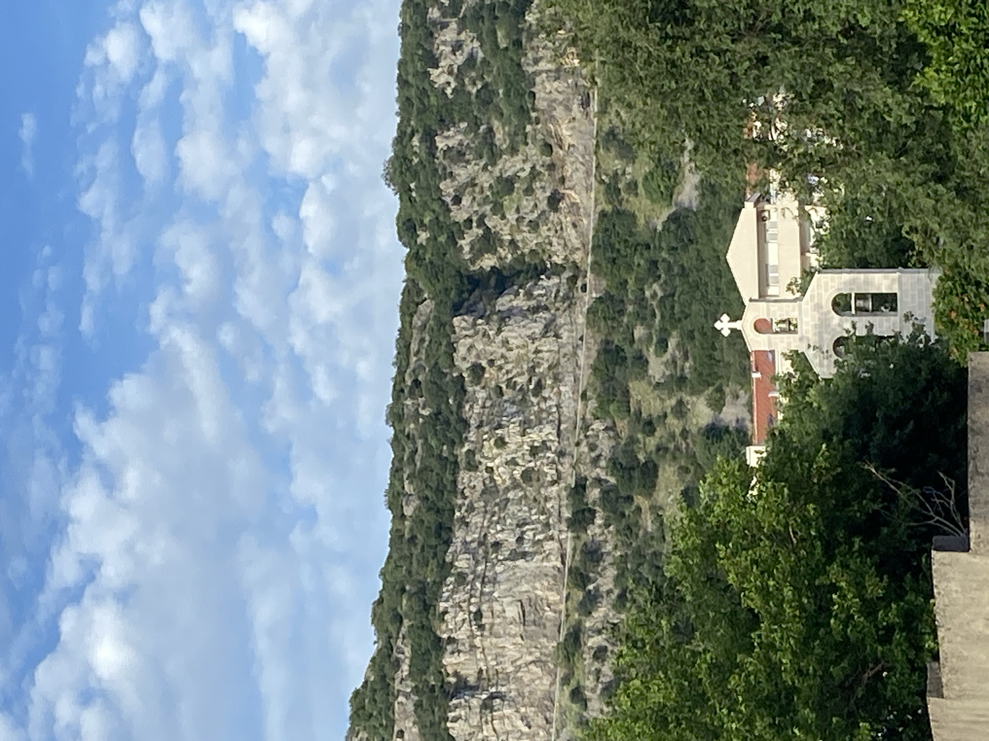 The image displays a mountainous coastal landscape under a bright blue sky with scattered white cumulus clouds. A prominent rocky mountain face in the background is heavily vegetated with green Mediterranean trees and shrubs, interspersed with exposed grey and tan rock formations. A dark asphalt road horizontally traverses the mid-slope of the mountain, winding through the terrain. In the mid-ground, a small, light-colored stone church is visible, featuring a bell tower with a cross on its apex and two arched openings housing bells. Behind and around the church, parts of other buildings are discernible, characterized by light-colored facades and reddish-brown tiled roofs. One multi-story building behind the church shows multiple windows and an external air conditioning unit. Dense green foliage and trees are present in the immediate foreground, partially obscuring a concrete or stone wall, and are also integrated throughout the mid-ground, surrounding the structures and extending up the mountain. No people are visible. The scene's architecture, vegetation, and mountainous coastal characteristics are consistent with the region of Budva, Montenegro.