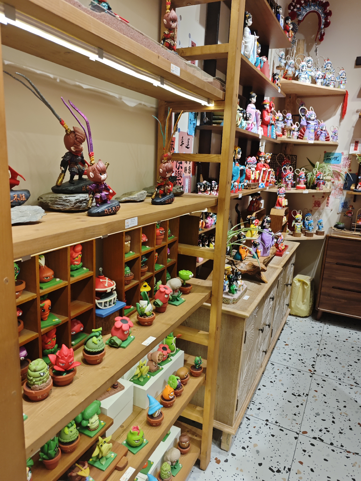 A display of figurines and other collectibles is visible in a store located in the Shapingba District, China. The shelves are wooden and hold a variety of items. The top shelf displays larger figurines of what appear to be mythological characters, while the shelves below showcase smaller figures that resemble cartoonish plants or vegetables in pots, each in its own cubicle. There is also a wooden ladder placed to the right of the left side shelves which leads to more shelves with traditional Chinese figurines. A wooden cabinet with floral designs on the door sits to the right of the shelves. The floor is tiled with a speckled pattern, and a yellow plastic bag is visible on the floor next to the cabinet. The scene is well-lit, with a clear view of the merchandise on display.