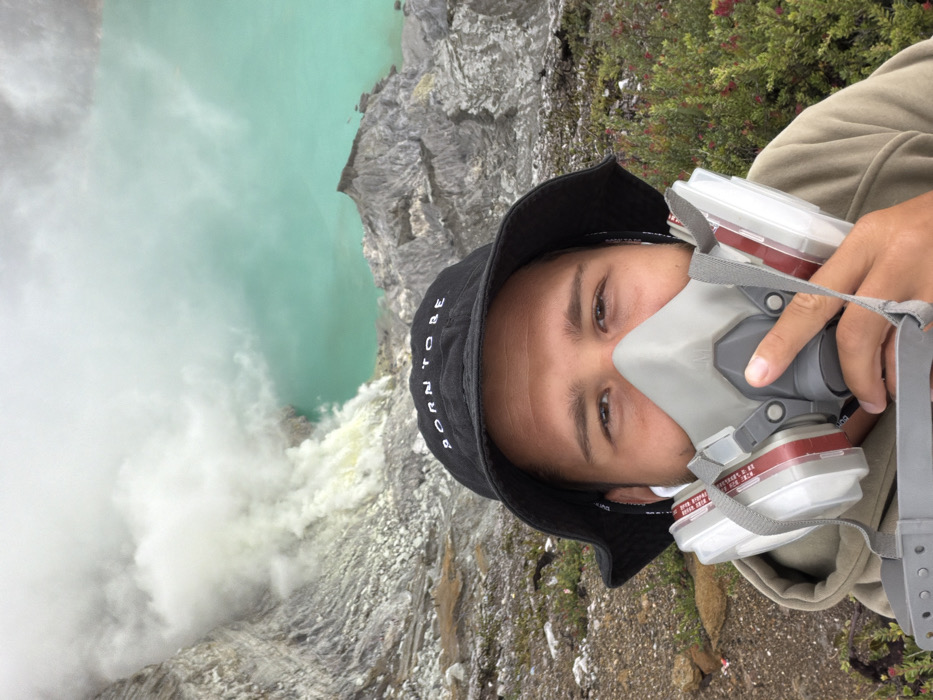 A person wearing a gas mask and a black bucket hat stands near a volcanic crater. The crater contains a pool of vibrant turquoise water, partially obscured by rising steam and mist. The surrounding terrain is rocky and barren, with some green vegetation visible. The person is holding the gas mask, likely protecting against volcanic gases. The image suggests an adventurous or hazardous environment.
