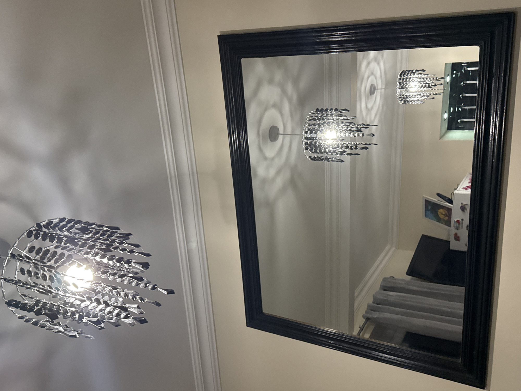 The image shows a room with light walls and white crown molding along the ceiling. A chandelier hangs from the ceiling. A large rectangular mirror with a black frame is mounted on the wall. The mirror reflects another chandelier, a narrow black storage unit, and a bed or sofa with gray textured fabric. The location in Gorleston-on-Sea, United Kingdom is unknown.