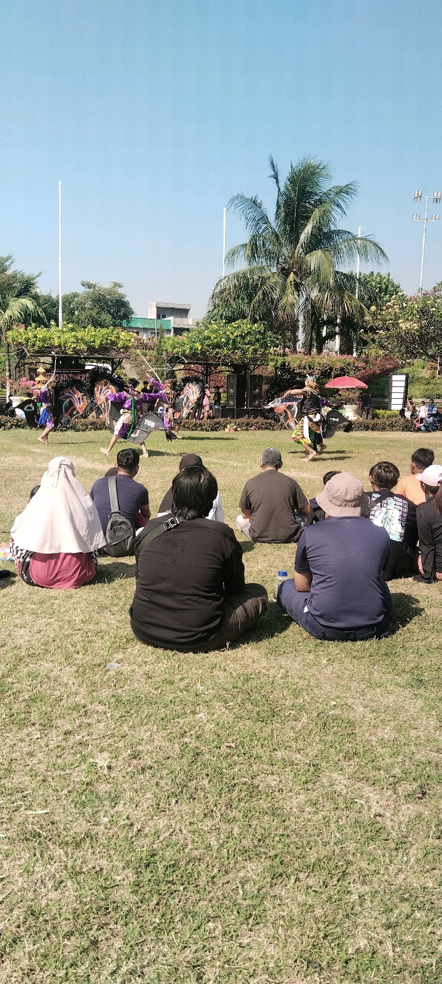 In Surabaya, Indonesia, a group of people are seated on a grassy field, facing a performance taking place in the distance. The performers, dressed in traditional vibrant costumes, are engaged in a dance routine, some holding large, decorative fans and staffs. Behind the performers, a large palm tree dominates the scene, with lush greenery and trees forming a backdrop. Buildings and speakers are visible in the background, suggesting an outdoor event. The sky is clear and blue, indicating a sunny day.