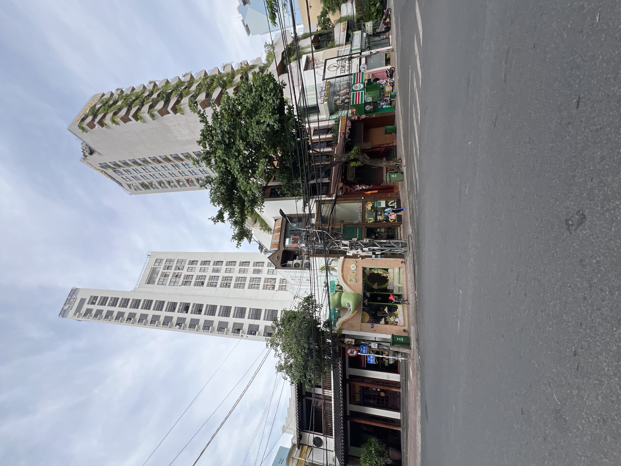 The image depicts a street scene in Nha Trang, Vietnam, featuring a mix of modern high-rise buildings and older, lower-rise establishments. In the background, two prominent white high-rise buildings dominate the skyline. The building on the left has a regular grid of windows and balconies, while the building on the right features a more striking design with cascading greenery on its balconies. In the foreground, several smaller buildings line the street. One of these has a distinctive green dinosaur sculpture on its facade, with the text "Dino's" above the entrance. Adjacent to it is a restaurant with traditional Vietnamese architecture, displaying signage that appears to read "Nam Phuong." The street itself is paved, with a pedestrian crossing marked by white stripes. Overhead, a dense network of electrical wires crisscrosses the sky. A few people are visible on the sidewalk, including one person sweeping. Several motorcycles are parked along the street. The sky is overcast with clouds.