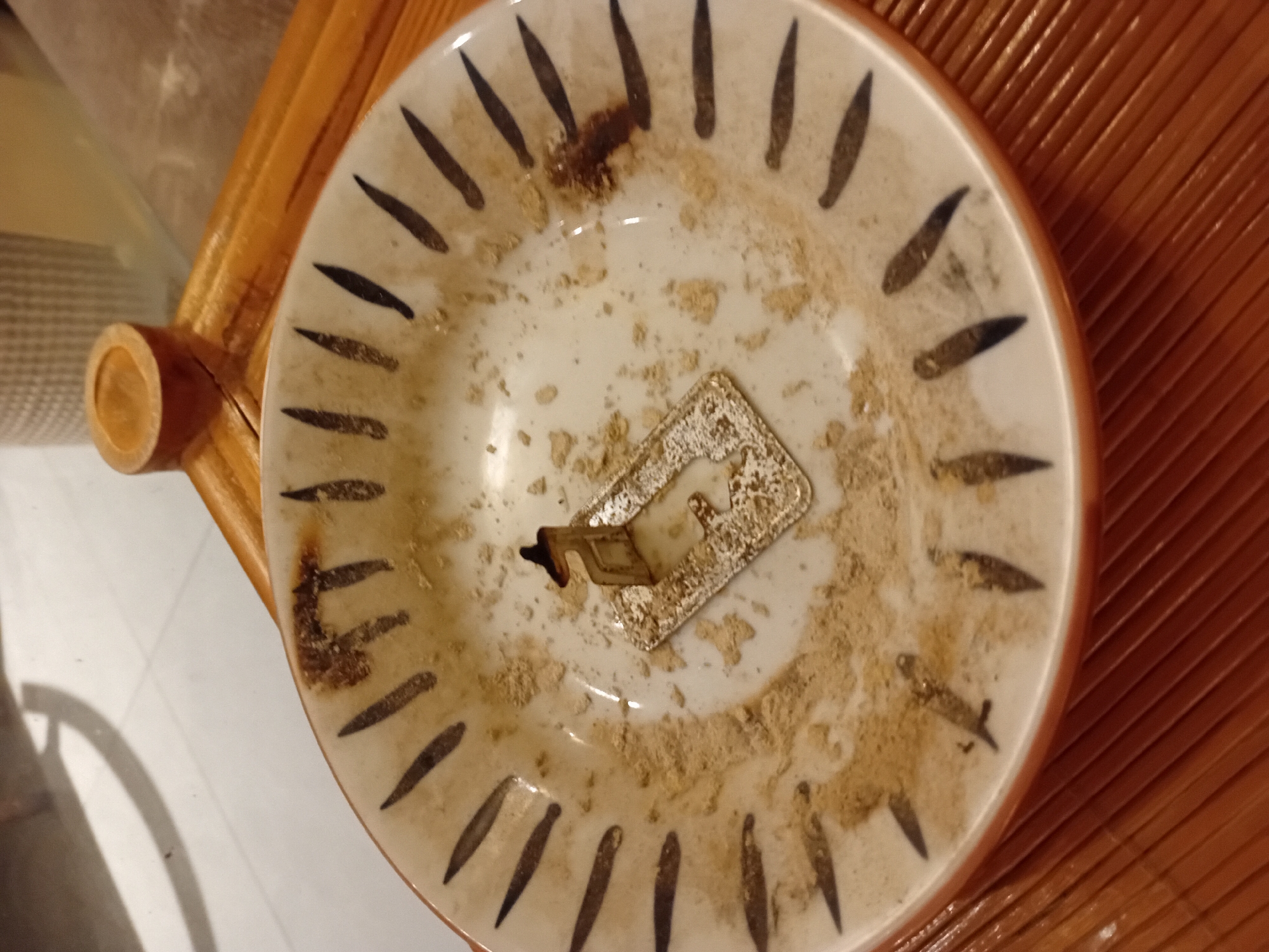 The image shows a close-up, top-down view of an ashtray resting on a woven wooden surface, possibly a chair or table, which is common in outdoor or semi-outdoor settings in Vietnam. The ashtray is made of ceramic and features a decorative pattern of dark, elongated strokes around its rim. The interior of the ashtray is filled with ash and cigarette butts. A metal stand, likely for resting cigarettes, is positioned in the center of the ashtray. The lighting suggests it is daytime, with shadows casting on the left side of the image, indicating a light source from the right. The overall scene is one of casual use, implying a moment of rest or a break. The location cues, such as the woven wood and the general aesthetic, are consistent with environments found in Đà Nẵng, Vietnam.