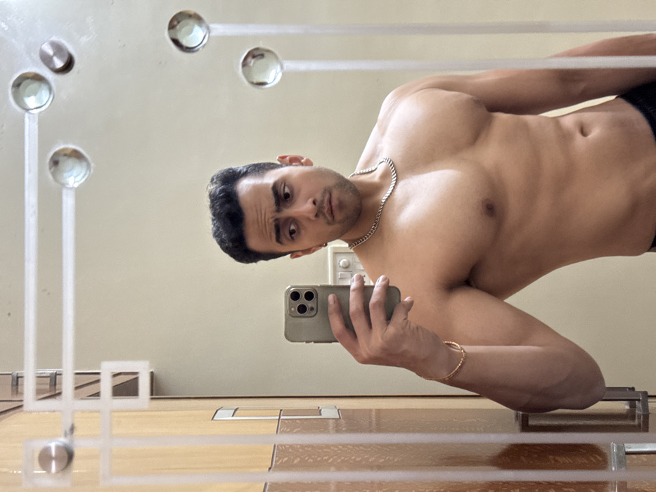 A shirtless man takes a selfie in a mirror, his head tilted to the left. He sports a silver chain around his neck and a gold bracelet on his wrist. The phone, gray with three visible camera lenses, is held in his left hand. In the background, a cream-colored wall is partially visible, along with light-colored floor tiles and decorative elements on the mirror itself. The man's facial expression is one of slight curiosity.