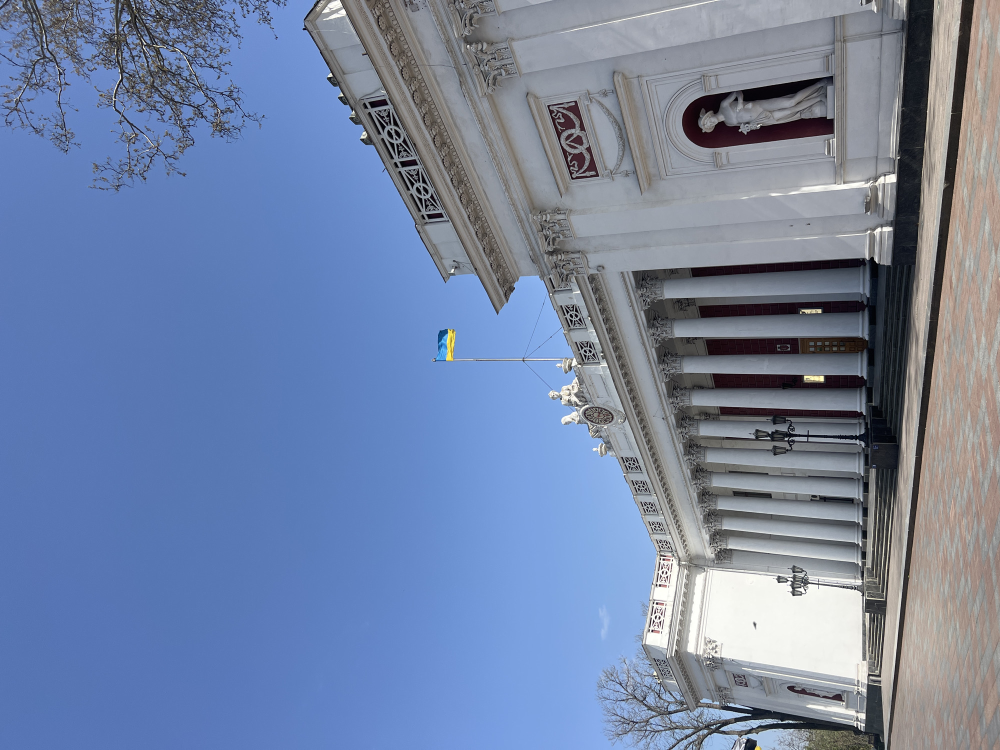 The image shows the Odessa Opera and Ballet Theater in Odesa, Ukraine. The white building features neoclassical architecture, including columns and a statue in a niche. A Ukrainian flag flies from a pole on the building's roof. The sky is blue and clear. Bare tree branches are visible on either side of the building. Part of a brick-paved walkway is visible on the right side of the image.