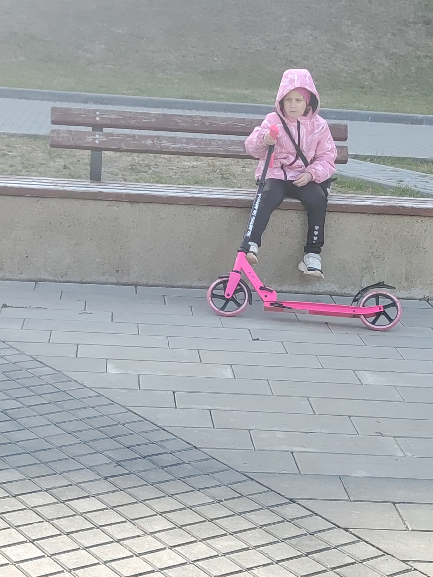 The image shows a young girl resting on a park bench next to her pink scooter in an outdoor setting in Chaikovsky, Russia. The girl, likely of school age, is seated on a wooden bench with a concrete base, her legs dangling. She is wearing a pink hooded jacket with a faint pattern, a pink beanie visible under the hood, black athletic pants with partially visible white text or design, and white sneakers. Her expression appears serious or pensive as she looks towards the viewer, holding onto the handle of her scooter.
The pink kick scooter lies on its side on the paved ground in front of the bench. It features black wheels with pink accents and a black fender. The ground is covered with grey paving stones, some rectangular and some square, laid in distinct patterns. Behind the bench, a grassy area is visible, with another paved path in the background. The lighting suggests it is daytime, possibly slightly hazy or overcast, as there are no strong, defined shadows. The scene depicts a quiet moment of rest or waiting during an outdoor activity.
