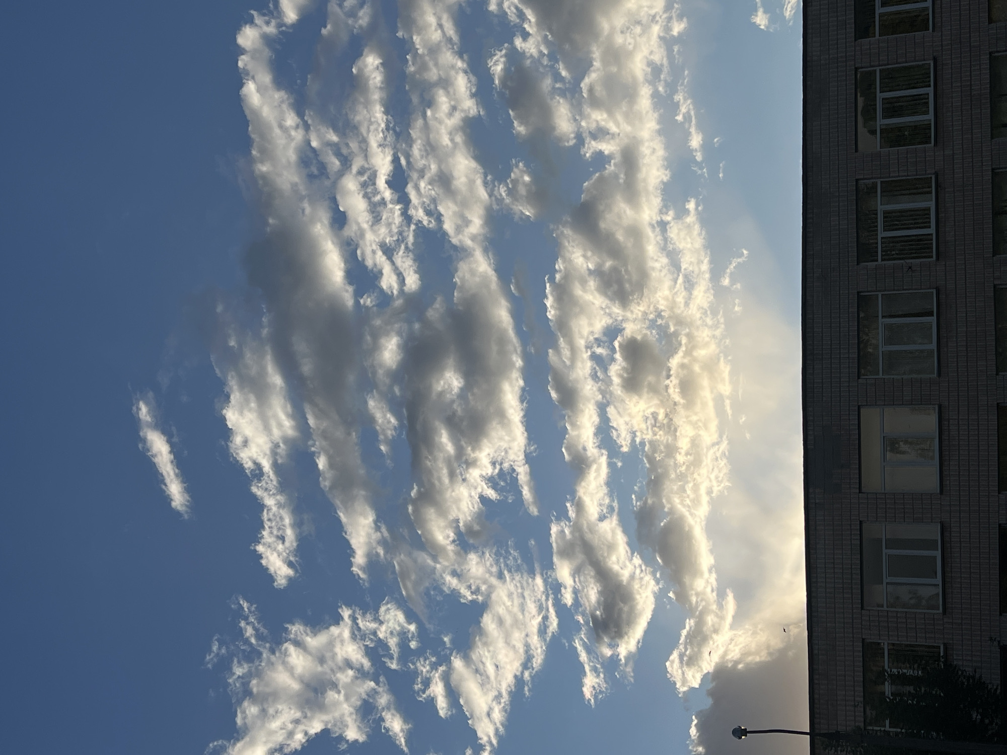 The image shows a view of the sky and a building in Ternopil, Ukraine. The sky is mostly blue with streaks of white clouds. The building is gray, with windows visible. A lamp post is visible at the bottom of the image.