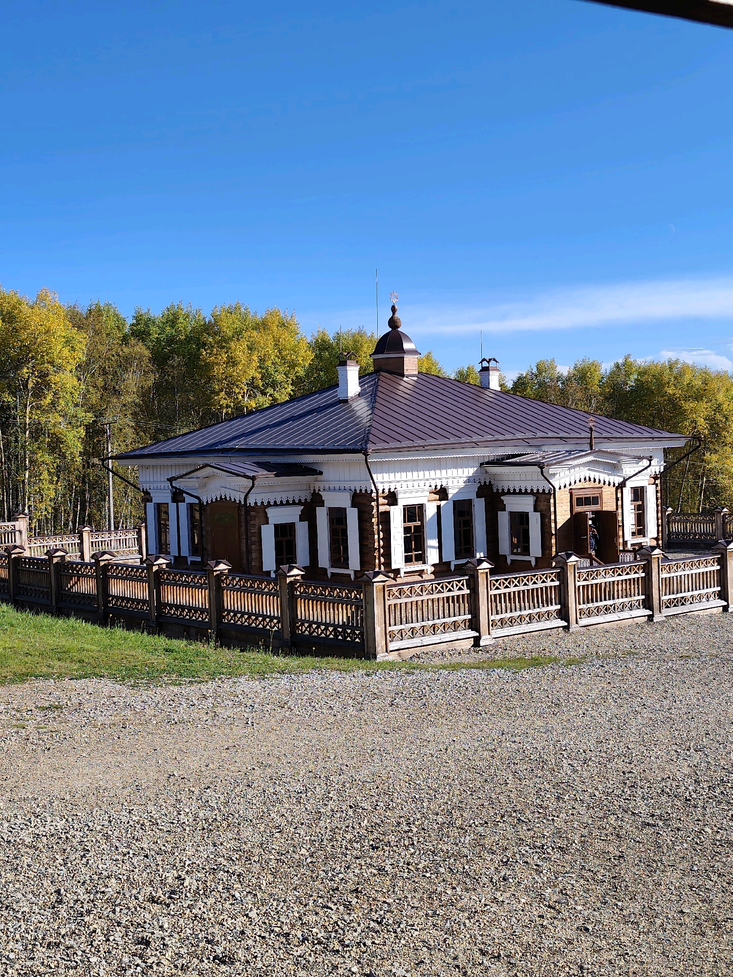 The image shows a traditional wooden building with a dark metal roof, likely a church or a historical dwelling, situated in a fenced enclosure. The building features intricate wooden carvings and white trim around the windows and eaves. A wooden fence, also decorated with lattice-like patterns, surrounds the property. Behind the building, a dense forest with autumn foliage, characterized by yellow and green leaves, creates a natural backdrop. The sky above is a clear, bright blue. The foreground consists of a gravel-covered ground. The location is identified as the museum of "Taltsy" in the Irkutsk region of Russia. There is one person visible near the entrance of the building, partially obscured.
