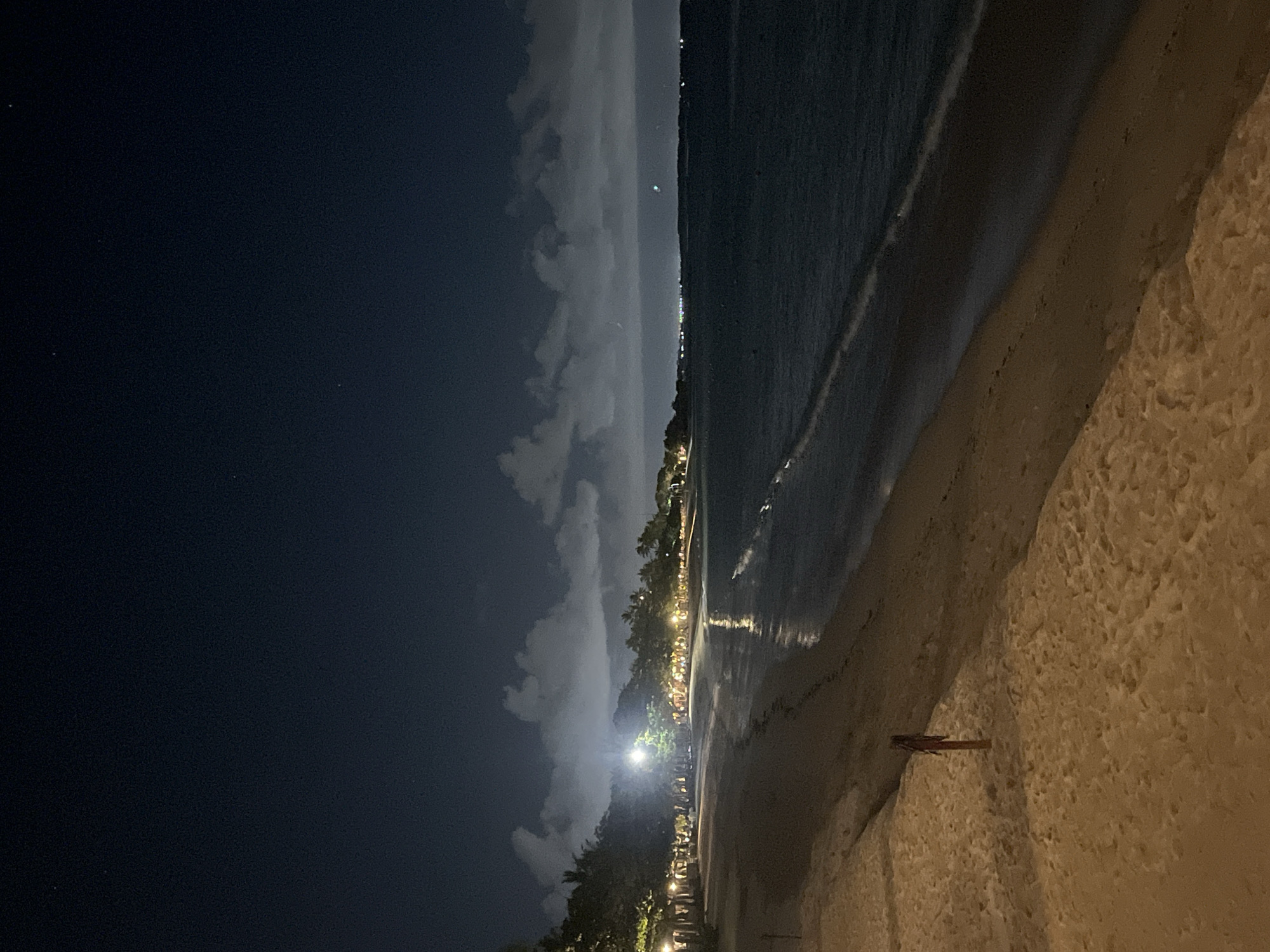 The image depicts a beach at night, likely in Kuta Selatan, Indonesia, based on the presence of a beach and what appears to be a tropical coastline illuminated by distant lights. The foreground shows a sandy beach, while the middle ground consists of the ocean's edge meeting the shore. A distant line of lights is visible along the coastline. The sky is dark, partially covered with clouds. A single figure, possibly an individual standing or sitting, is visible on the beach to the right of the image.