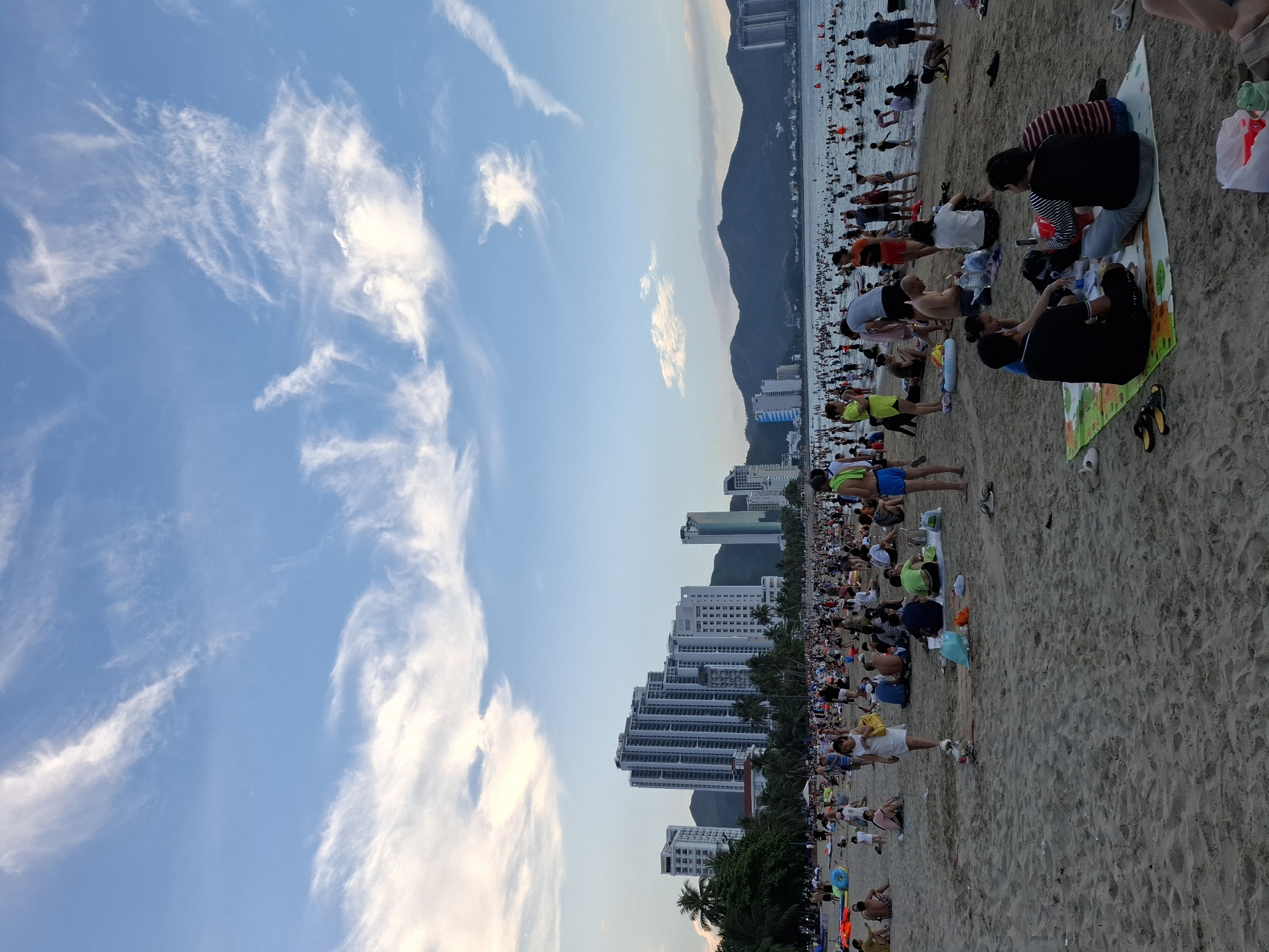 The image depicts a crowded beach in Nha Trang, Vietnam. The foreground shows a sandy beach with several groups of people sitting on blankets and relaxing. Some individuals are standing and walking around. The middle ground is occupied by a large number of people wading and swimming in the ocean. In the background, there are numerous high-rise buildings lining the coast, characteristic of a city. Mountains are visible behind the buildings, and the sky is partly cloudy with wispy cirrus clouds. The overall scene suggests a popular tourist destination during daylight hours, likely late afternoon or early evening given the lighting.