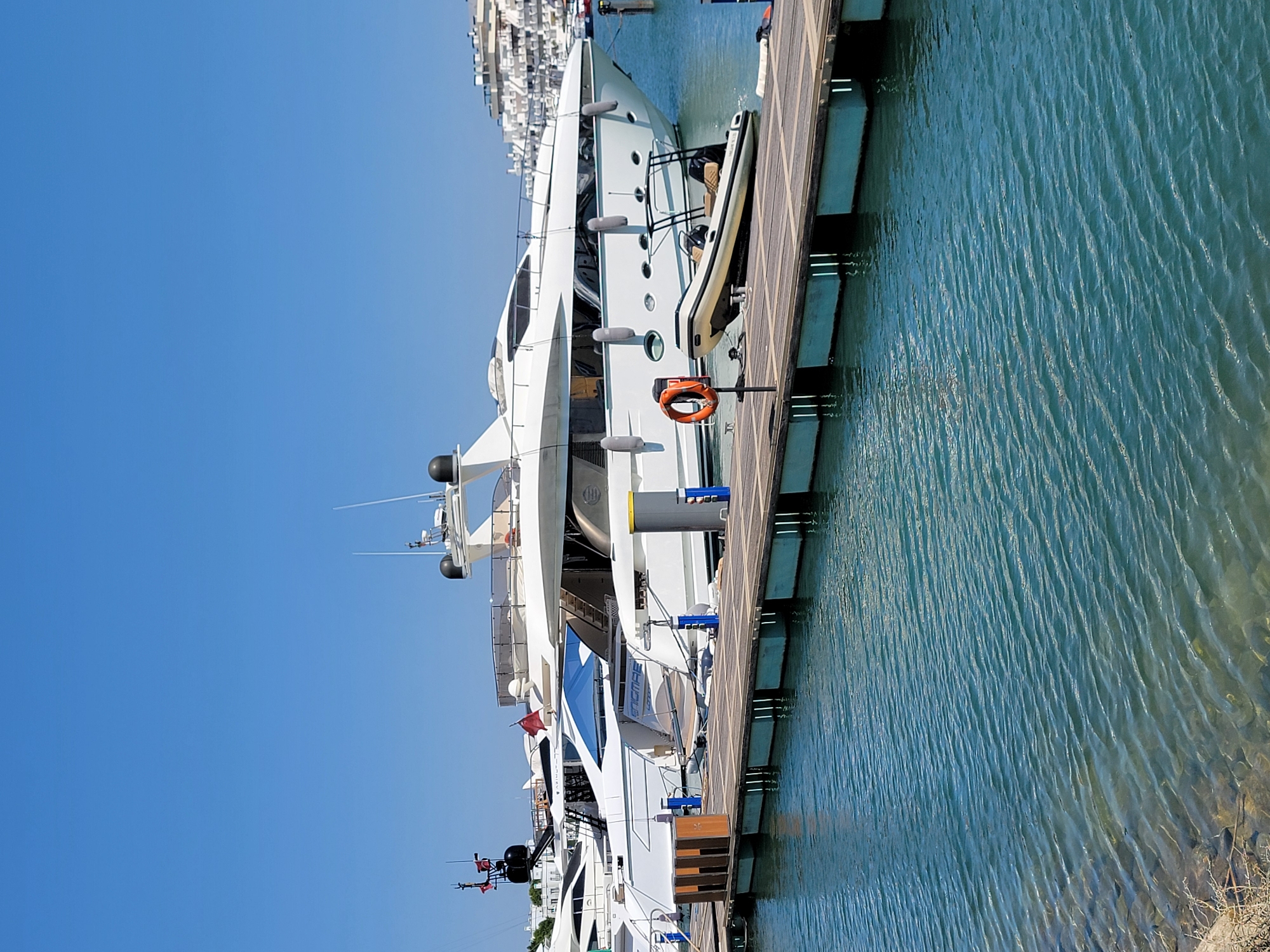 In the city of Quarteira, Portugal, a large, white luxury yacht is docked at a wooden pier on a clear, sunny day. The yacht features multiple levels, sleek windows, and various antenna structures on its upper deck. A smaller inflatable boat is positioned on the pier next to the yacht, with its bow facing the water. An orange life preserver hangs on a post on the pier. The water is calm with gentle ripples. In the background, other buildings and structures, possibly more yachts or apartments, are visible along the waterfront. The sky is a vibrant blue.