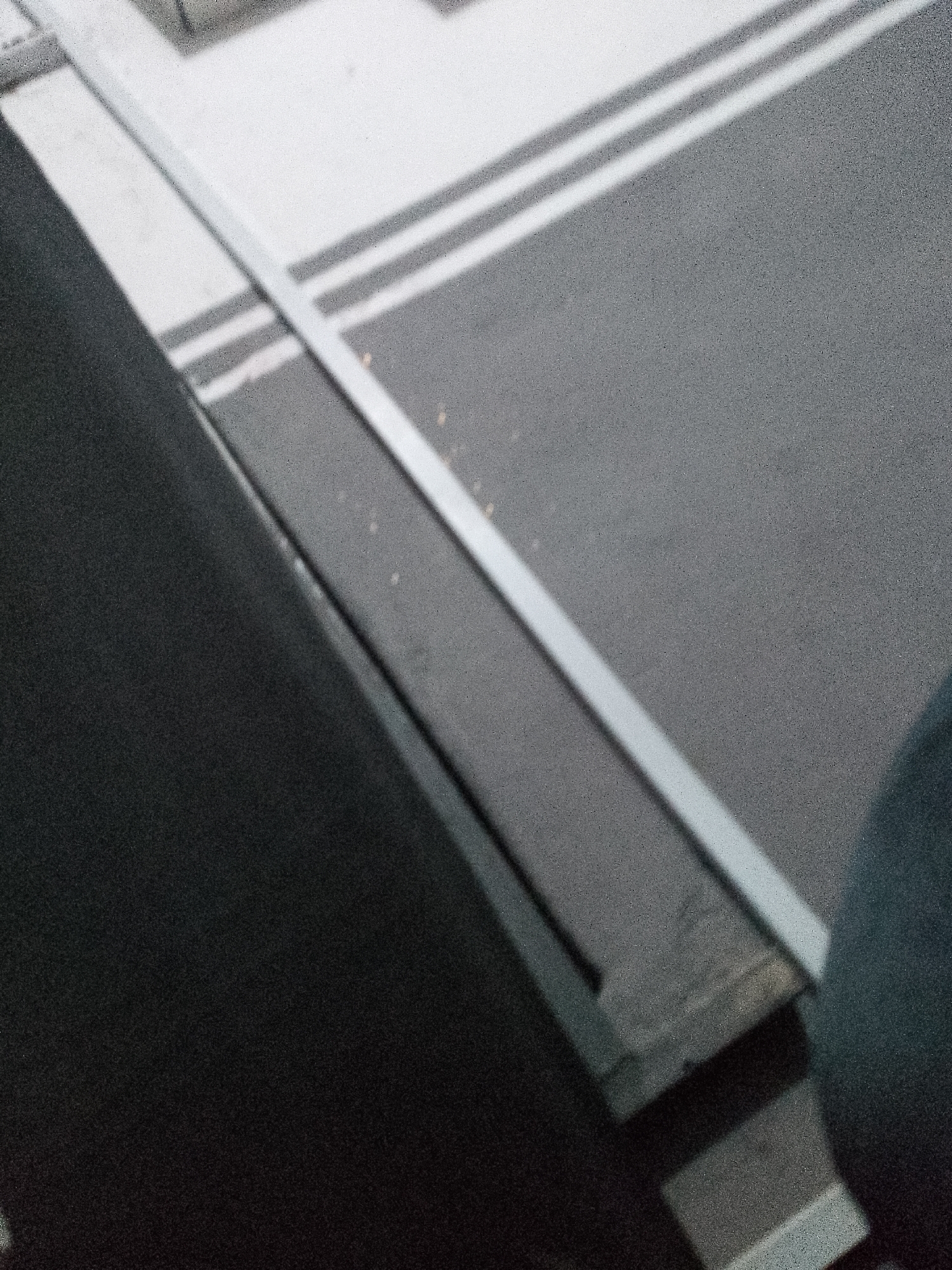 A close-up, grainy, over-the-shoulder view shows a dark, textured surface in the foreground. This surface appears to be a dark fabric, possibly clothing or upholstery, angled diagonally from the bottom left to the top right. Beyond this dark surface, a light-colored, metallic railing or bar runs diagonally across the image from the top left towards the bottom right. Behind the railing, a gray asphalt surface is visible, marked with two parallel white lines, suggesting a road or parking area. The lighting suggests an outdoor environment, potentially during daylight hours, as indicated by the presence of shadows cast by the railing. The image's perspective and lack of identifiable landmarks make it impossible to determine the specific location in Jalingo, Nigeria.