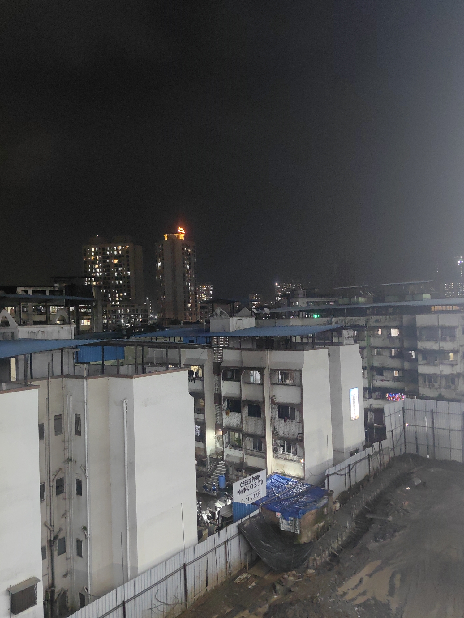 The image depicts a nighttime view of a densely populated urban area in Mira-Bhayander, India. The foreground features the tops of several buildings with white facades and blue corrugated metal roofs. A construction site is visible on the right, with a metal fence, dirt, and debris. In the middle ground, there are more apartment buildings with many illuminated windows, suggesting residents are present. A sign on one building reads "GREEN PARK MAHAL CHS LTD MADAR." Further in the background, taller buildings are visible, with one illuminated by a neon sign. The sky is dark, indicating it is nighttime, and the overall scene suggests a typical urban environment with residential and possibly commercial structures. There are no visible individuals in the frame.