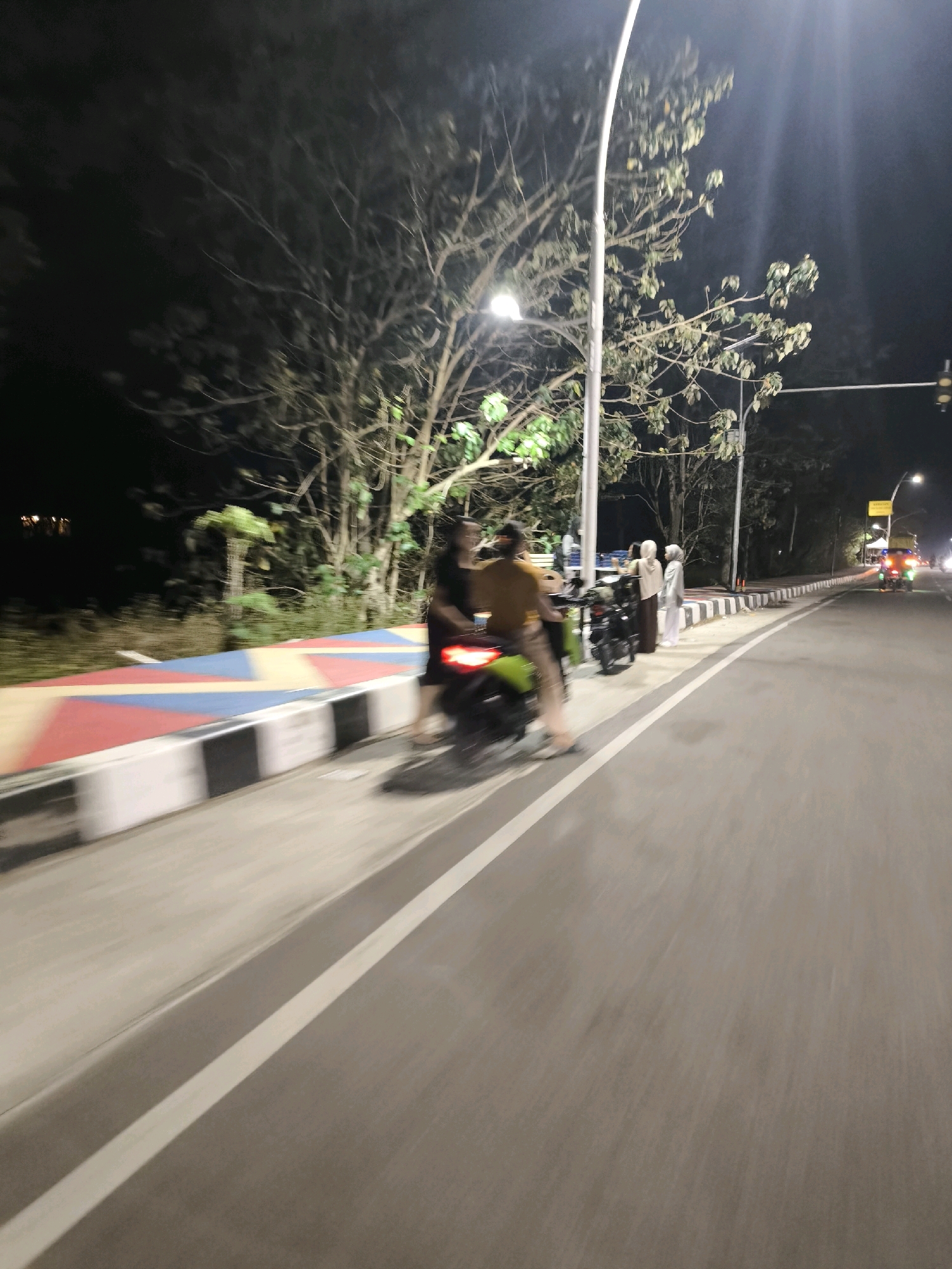 The image depicts a nighttime scene on a street in Kwandang, Indonesia. The foreground shows a portion of a road with a white lane marker. To the left, a sidewalk area is visible with a decorative barrier featuring red, yellow, and blue triangular patterns, bordered by black and white striped sections. Several people are on or near the sidewalk, some standing and others on a green motorcycle. One individual in a yellow shirt is seated on the motorcycle, which is blurred due to motion. Other individuals, appearing to be women wearing hijabs, are standing near parked motorcycles. In the background, trees line the side of the road, illuminated by streetlights. Further down the road, traffic lights and other vehicles are visible, suggesting active traffic. The overall impression is that of a public street at night, possibly a commercial or recreational area, with people engaging in activities near the roadside.