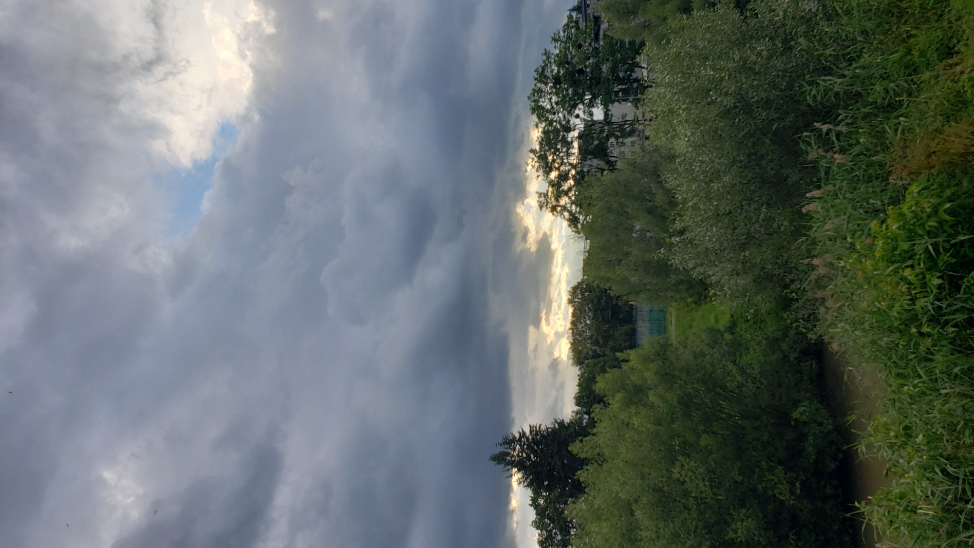 The image depicts a cloudy sky over a lush green landscape with trees and vegetation. In the distance, faint outlines of buildings are visible. A narrow body of water, possibly a river or stream, runs through the foreground, bordered by dense green reeds and foliage. The clouds are heavy and dark, with patches of lighter sky and sunlight breaking through in the upper right portion of the frame. A few small, dark shapes, likely birds, are visible against the sky. The overall impression is one of a natural setting with signs of urban development in the background. The location is identified as Prešov, Slovakia.