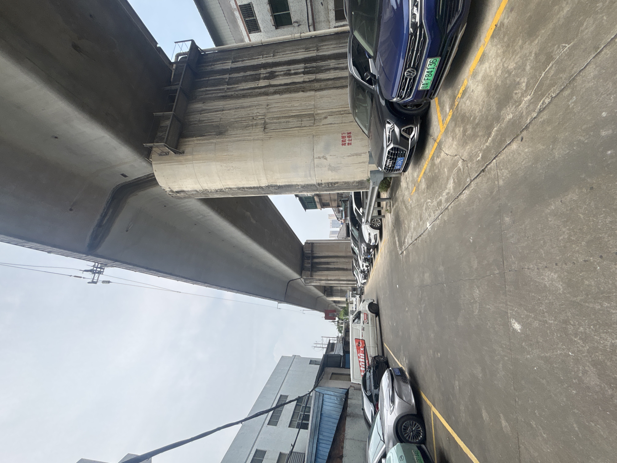 An exterior, eye-level, medium shot captures a parking lot beneath an elevated highway in Nanhai District, China. The concrete overpass dominates the upper portion of the frame, supported by massive, weathered concrete pillars. The sky is overcast and pale. Several cars are parked in designated spots marked by yellow lines. In the foreground, to the left, a silver sedan is partially visible, followed by a black sedan. Further down the row of parked cars, an Audi and a blue Volkswagen are prominent. A white van with red lettering is parked in the mid-ground. The ground is asphalt, cracked and stained. Buildings are visible in the background, including a white industrial-style building to the left and other structures further away. Red Chinese characters are visible on one of the concrete pillars. The scene appears to be a typical urban parking area with a focus on the infrastructure of the elevated highway.