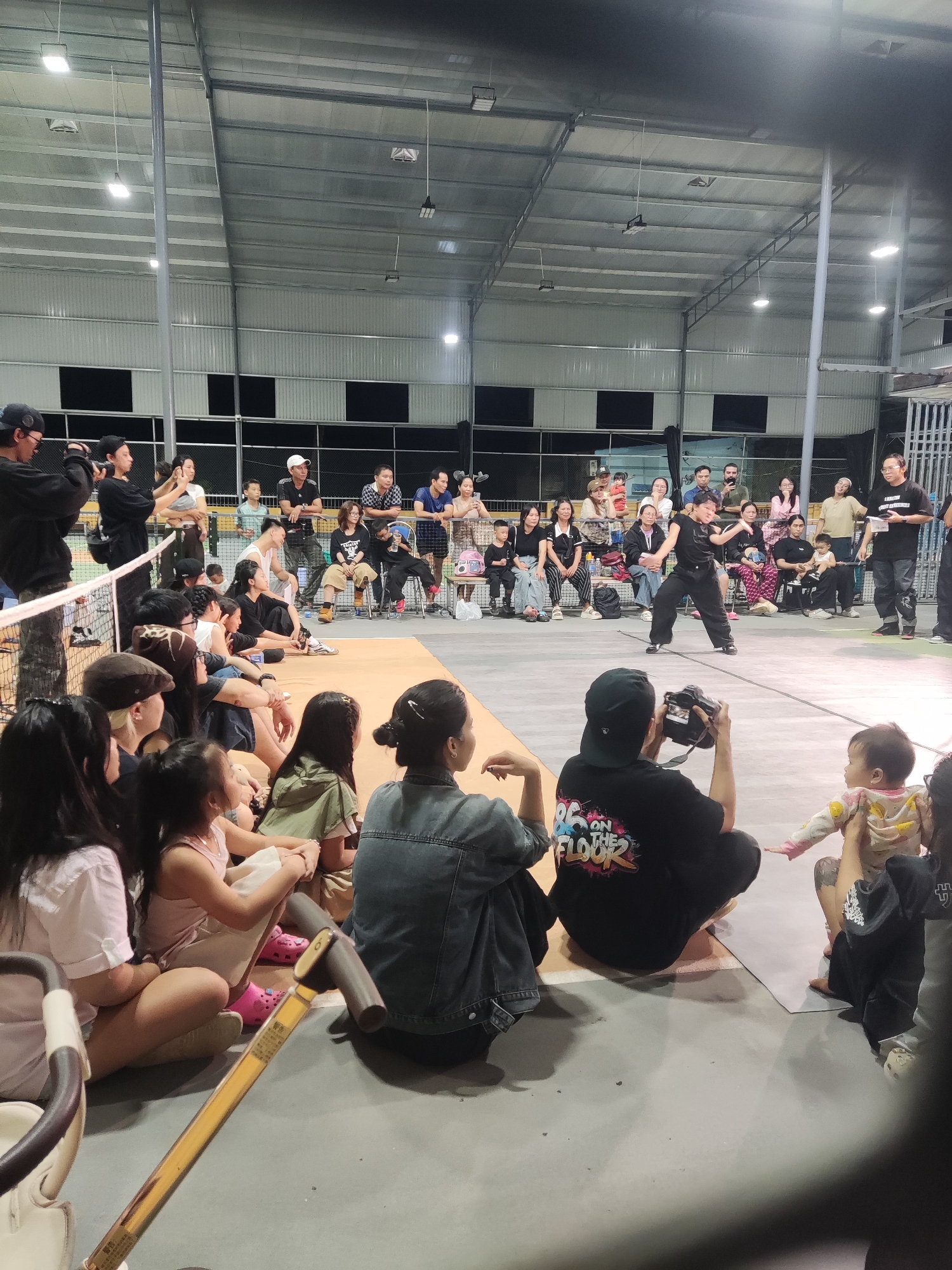 A performance is taking place in a large, indoor arena with a metal roof and support beams. The audience, composed of adults and children, is seated on the floor or on chairs along the perimeter. A young adult in black clothing is dancing in the center, with photographers capturing the performance. The arena is well-lit with overhead lighting fixtures. A tennis net is visible in the foreground.