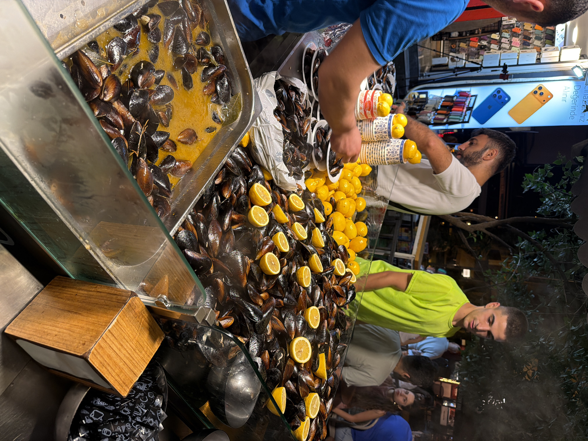 The image depicts a nighttime scene at a street food stall in Kadıköy, Turkey, specializing in mussels. A man in a blue shirt is seen serving mussels from a large metal tray filled with a golden broth and mussels. In the foreground, a large display case is filled with freshly cooked mussels, garnished with numerous lemon slices. To the left, stacked containers with lemons are visible, suggesting they are for serving with the mussels. Behind the mussels, a large white bag is filled with more mussels. In the background, a man in a bright green shirt stands next to the display, looking down. Other people are visible further back, appearing to be customers. A brightly lit sign showcasing phone cases is on the left side of the frame, and various street elements indicate an urban setting. A wooden napkin dispenser is placed on the counter in the lower right quadrant of the image.