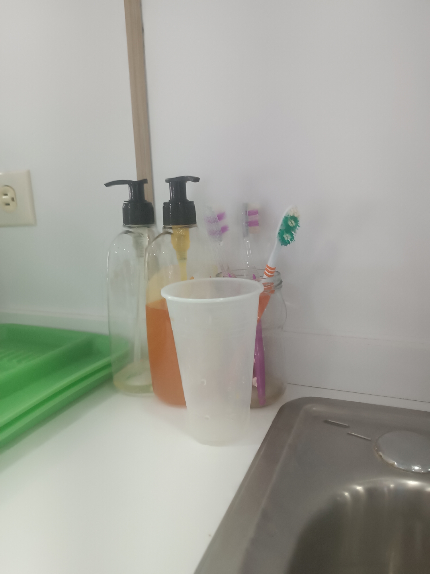 The image shows a bathroom counter with various items, including two pump bottles of liquid soap, one empty and one containing orange liquid. Next to them, a white plastic cup and a glass jar holding several toothbrushes are visible. A green tray is placed to the left of the pump bottles, and a sink with a drain is in the foreground. The scene is set in a bathroom in Porlamar, Venezuela.