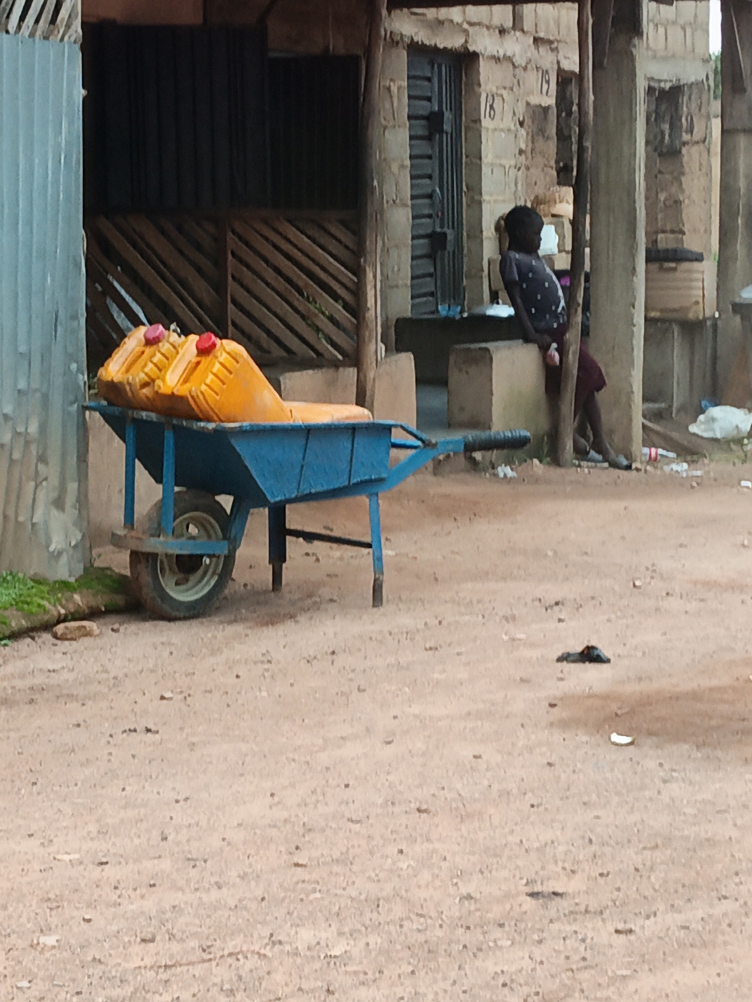 In an outdoor, urban setting, a blue wheelbarrow is positioned on a dirt ground, carrying three orange plastic jerrycans with red caps. The jerrycans are stacked on top of each other, with two in the front and one behind. The wheelbarrow has a single wheel and metal handles. In the background, a person wearing a dark shirt with white polka dots and a maroon skirt is seated, leaning against a concrete pillar. They are holding a light-colored object in their right hand. Behind the person is a building constructed of concrete blocks. The ground is mostly dirt with scattered debris, including some white plastic bottles and a dark, flat object that could be a shoe. The environment appears to be an undeveloped or partially developed area, possibly a market or a residential street.