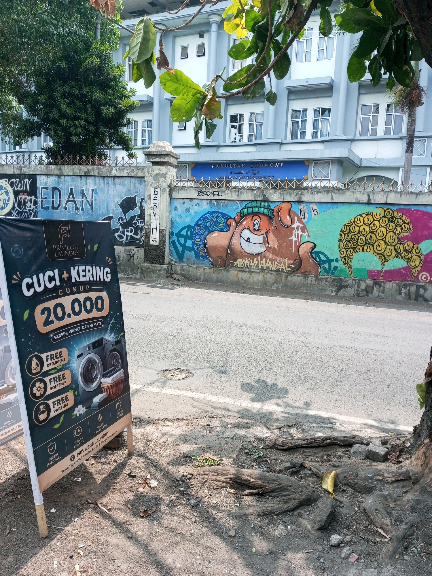 This is a street-level, medium-shot photograph taken on a sunny day in the city of Malang, Indonesia. In the foreground, a black A-frame sign for "Privilege Laundry" is propped up by wooden sticks on a gravelly sidewalk. The sign advertises "CUCI + KERING CUKUP 20.000" (Wash + Dry Enough 20,000) and lists "BERSIH, WANGI, DAN HEMAT" (Clean, Fragrant, and Economical). It also mentions "FREE DETERGENT", "FREE SOFTENER", and "FREE PARFUM". Below these offers, there are icons representing speed, practicality, and maximum cleanliness.
Behind the sign, a gray asphalt road stretches into the distance. In the mid-ground, a concrete wall is covered in graffiti and murals. One mural features a cartoonish, brown, pig-like character with a green beanie, while another depicts a yellow mass of smiley faces. Above the wall, a light blue building with multiple windows is visible, identified by a sign reading "FAKULTAS EKONOMI" (Faculty of Economics). Lush green trees partially obscure the view of the building and the sky. The environment appears urban, with elements of street art and commercial advertising. The roots of a large tree are visible on the right side of the frame, encroaching on the sidewalk.