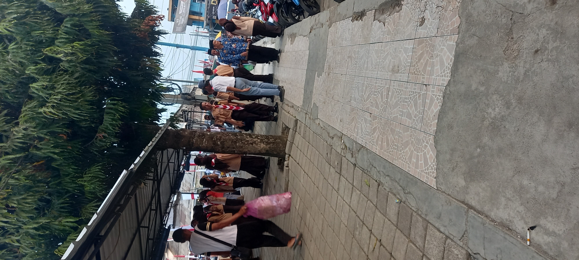 The image depicts an outdoor scene in Wanareja, Indonesia, likely near a school or community gathering, as evidenced by the presence of individuals in scout uniforms. The foreground shows a paved walkway constructed with two types of tiling: a coarser, gray concrete in the immediate foreground, and a lighter, patterned tile further back. A discarded cigarette is visible on the gray concrete.
Dominating the midground is a large tree with dense, green foliage, partially obscuring a covered walkway or awning. Several people are gathered under this awning and along the sidewalk. Many appear to be young people in scout uniforms, which consist of tan shirts, brown pants or skirts, and red and white neckerchiefs. Some adults are also present, including a man in a white shirt and blue pants, and another in a patterned blue shirt and dark pants. One individual, walking away from the viewer on the left, carries a large, translucent pink plastic bag.
In the background, there are buildings and what appear to be motorcycles parked along the edge of the scene, suggesting a public area or marketplace. The overall atmosphere seems to be one of a casual gathering or event.