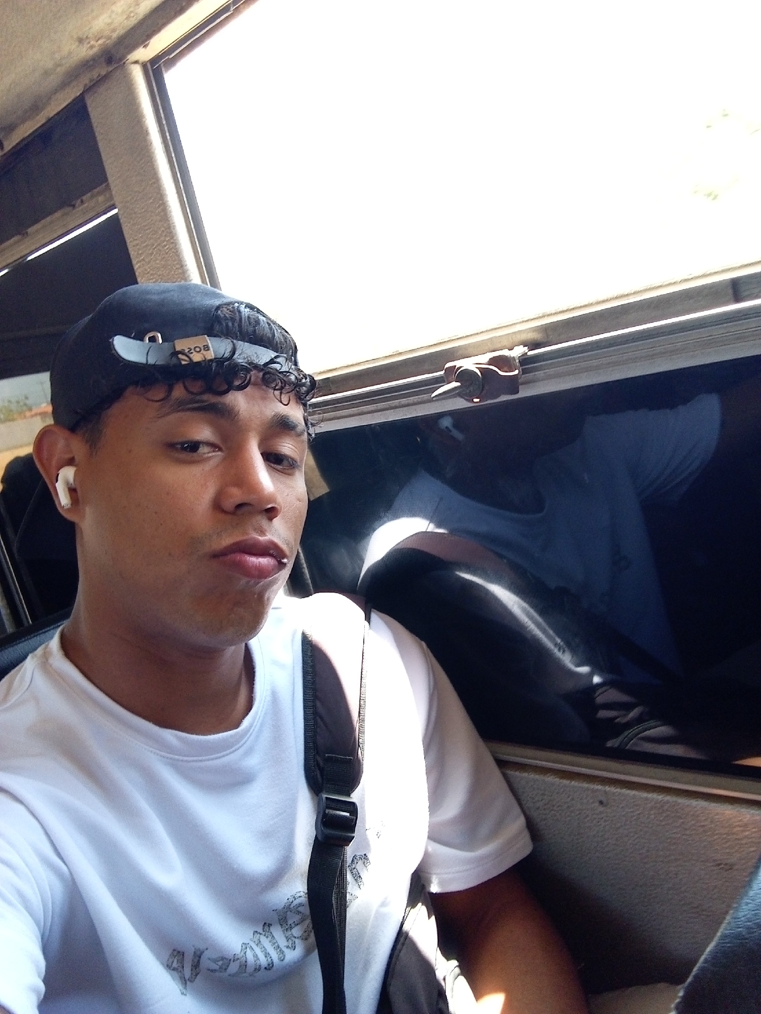 A young man is captured in a close-up, chest-up portrait inside what appears to be a vehicle. He has brown skin, short curly dark hair, and is wearing a black baseball cap backward, allowing some curls to peek out from under the brim. The adjustable strap on the back of his cap, which is light blue, clearly displays the word "BOSS" in dark lettering, appearing upside down due to the cap's orientation. He wears a plain white t-shirt with some faded, distressed gray text on the chest, which is largely illegible but might spell out "GAMES". A black strap from a backpack is visible over his right shoulder. He has a white earbud in his left ear.
The man is looking directly at the camera with a neutral expression, his lips slightly parted. The scene is brightly lit by natural sunlight streaming in through a large window on his right side, causing the background outside the window to be overexposed to white. The dark interior of the vehicle is visible around him, and a reflection of a person wearing a light-colored t-shirt can be seen in the window glass, suggesting another passenger or the subject's own reflection. The strong sunlight indicates it is daytime, likely a sunny day. The location appears to be inside a moving or stationary vehicle, possibly a bus or car.
