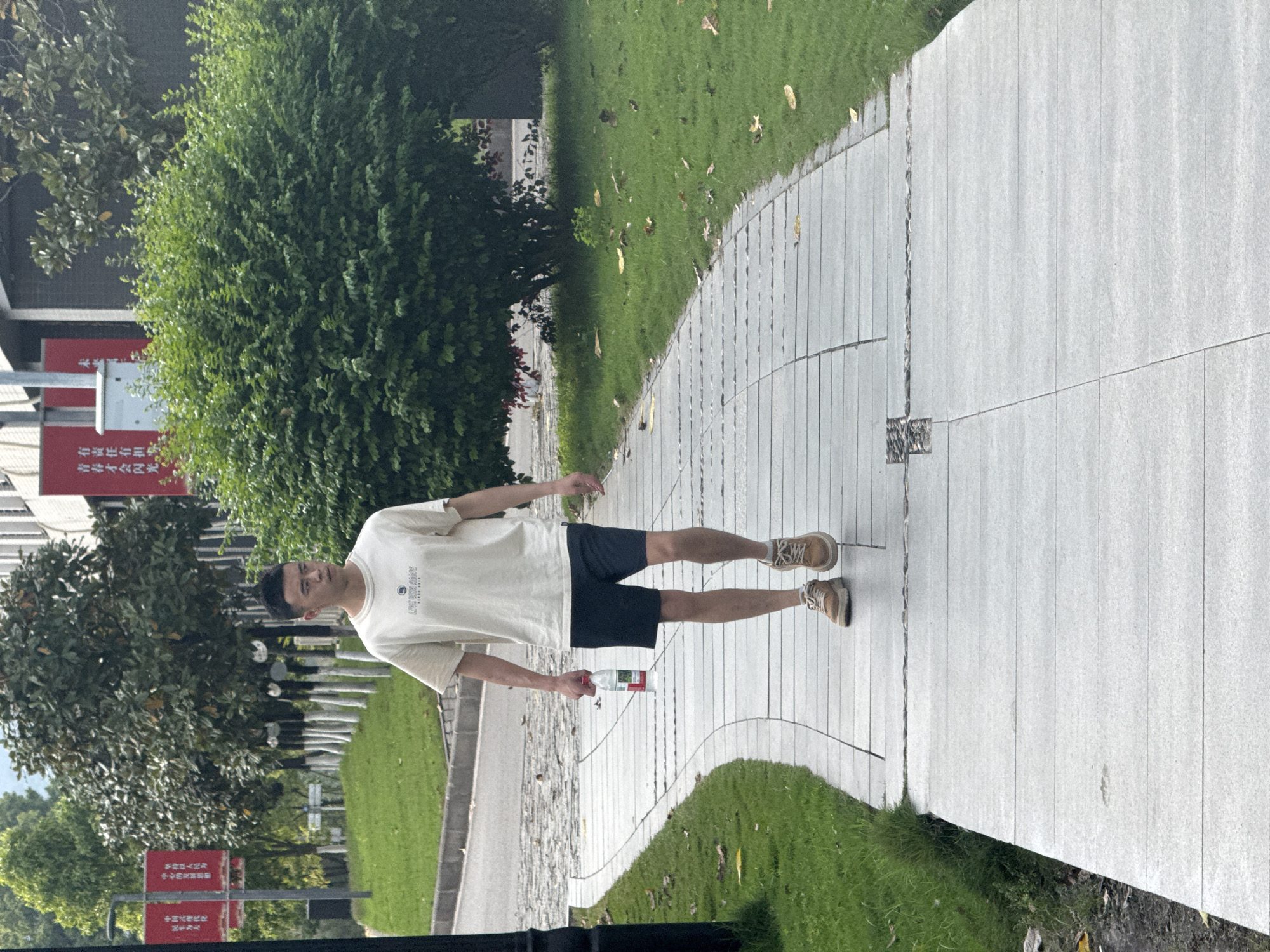 A young man is standing on a sidewalk in what appears to be a park or recreational area in Jiangjin, China. He is wearing a light-colored t-shirt, dark shorts, and tan sneakers. He is holding a bottle of water. The sidewalk is made of light gray, long, rectangular stones and borders a green lawn area. The background includes trees, greenery, and signage with Chinese characters, as well as an advertisement for "LIVE WITH AGOPS". A sign near the man reads "有责任有担 青春才会闪光".