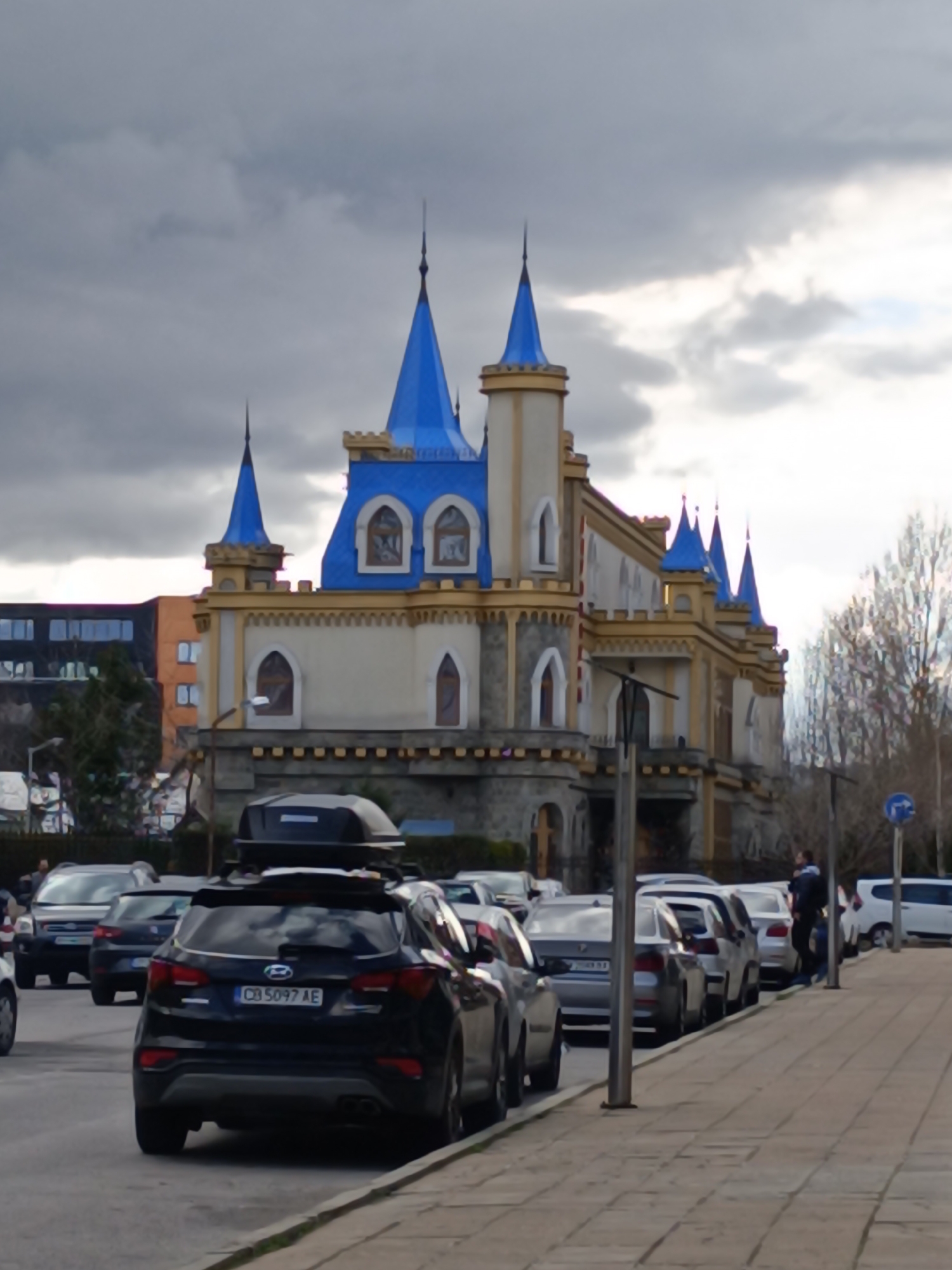 The image shows a street scene in Sofia, Bulgaria, with a fanciful castle-like building in the background, likely a restaurant or entertainment venue. The building is characterized by a light beige color scheme with blue turrets and gold trim. The sky is overcast.
In the foreground, a row of parked cars lines the street. A black SUV with a cargo carrier on top is prominently positioned. The car's license plate is visible, reading "CB 5097 AE." A person is standing near the sidewalk among the parked cars. Other vehicles and pedestrians are visible in the distance. The street is paved with patterned stone tiles. A street sign is also visible near the sidewalk. In the background, there are more buildings and bare trees.