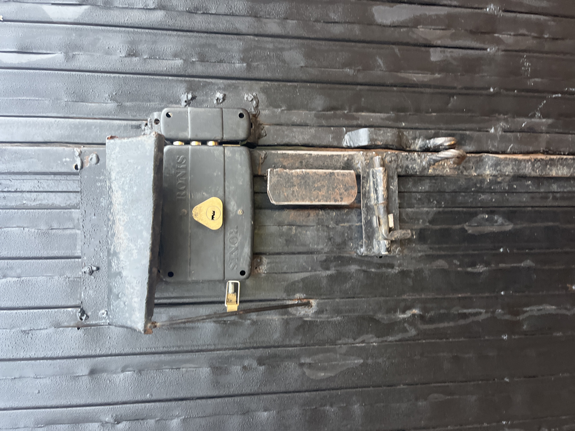 The image shows a close-up of a dark, metal, corrugated gate with a multi-point locking mechanism. The lock is centrally located and appears to be a heavy-duty, industrial-style lock. It has a gold-colored keyhole with a triangular escutcheon plate. Above the lock, there is a metal cover or shield, also dark and weathered. The gate itself is composed of vertical, ribbed metal panels, painted black. The surface of the gate shows signs of wear and tear, with some rust and water marks visible. The overall impression is one of security and durability. The image does not contain any people or evidence of recent activity. The scene is static and focused on the locking mechanism of the gate.