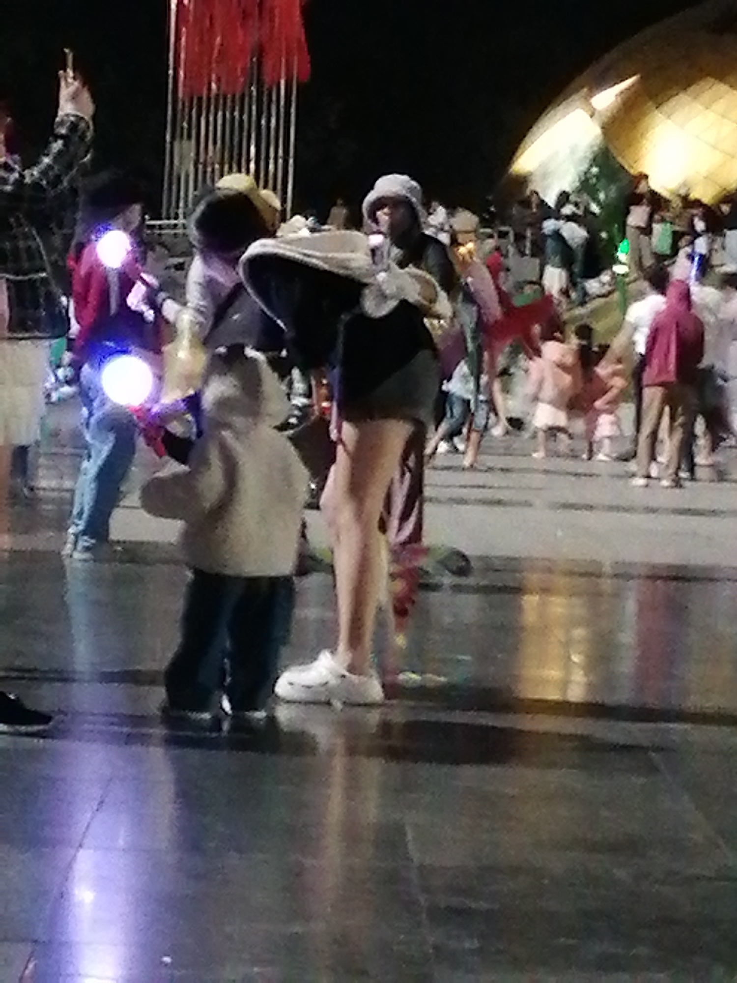The image captures a nighttime scene in Da Lat, Vietnam, likely at the night market given the presence of various stalls and a crowd of people. In the foreground, a young child wearing a light-colored hooded jacket and dark pants is holding a glowing blue orb object, possibly a toy. To the left of the child, an adult in a red jacket and blue jeans also holds a similar glowing object. A woman in a white hat and black clothing stands near the child, her left arm extended. Behind them, other individuals are visible, some browsing at stalls. The environment appears to be an outdoor public space with paved ground reflecting the ambient lights from the stalls and decorations, including red banners hanging above. In the background, a large, curved, golden-domed structure is illuminated. The overall atmosphere suggests a lively evening gathering or event.