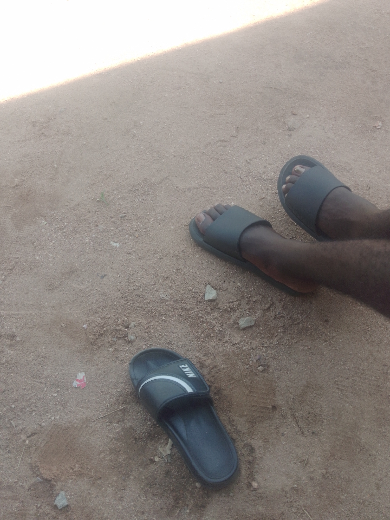 Two feet wearing dark colored slide sandals are visible on a sandy ground. One foot is pointing to the right, and the other is pointing towards the left. A black slide sandal with a white Nike logo is also visible on the ground to the left of the feet. The ground is a mixture of sand and small pebbles. A diagonal shadow is cast across the upper left portion of the image.