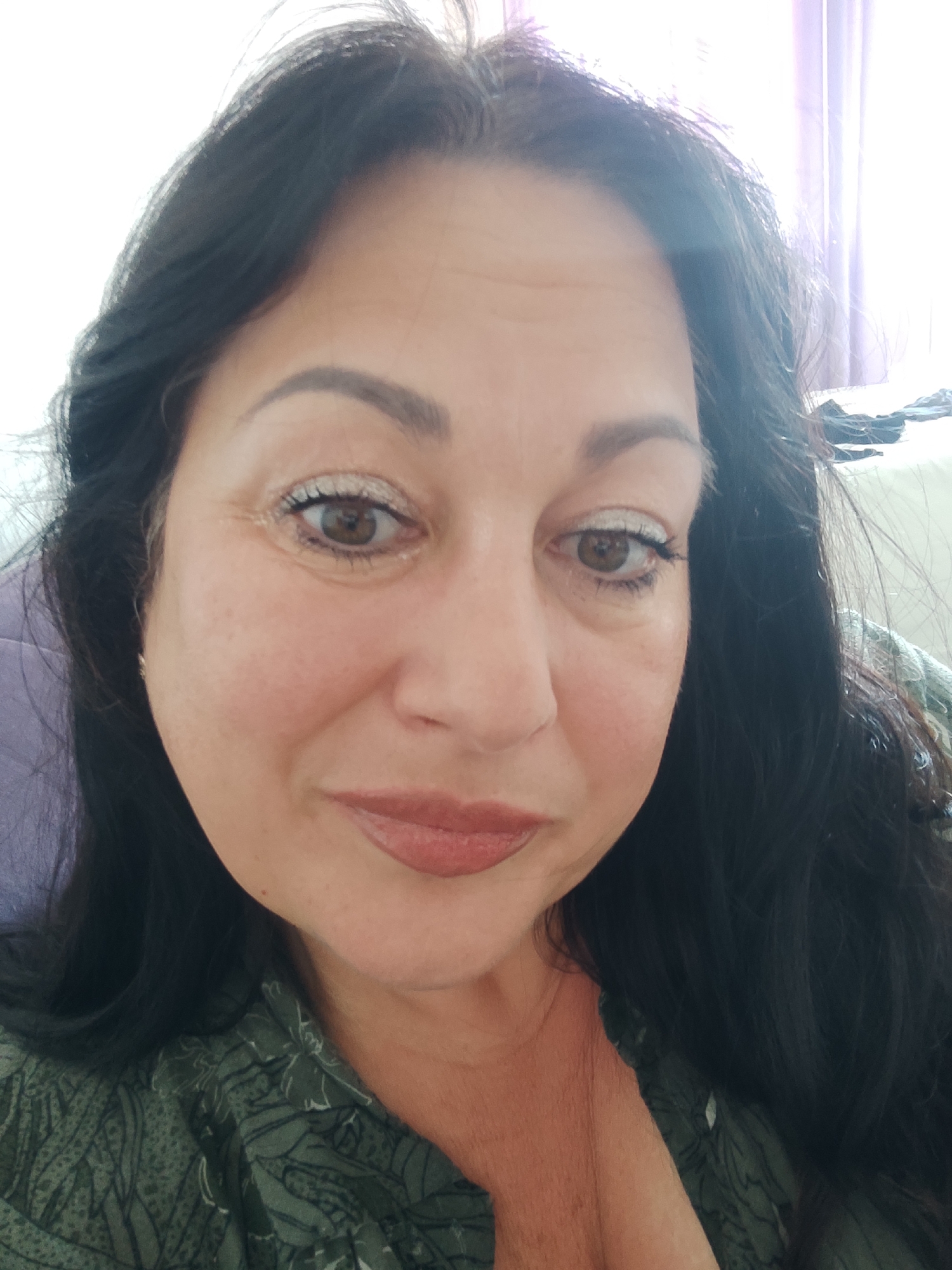 The image is a close-up, selfie-style portrait of a woman with dark hair, brown eyes, and fair skin. She is wearing makeup, including eyeshadow and lipstick. Her clothing appears to be a green patterned blouse. The background is out of focus, but suggests an indoor setting with natural light coming from a window. There are purple curtains visible. There is no indication of the city of Sochi, Russia, or any specific location.