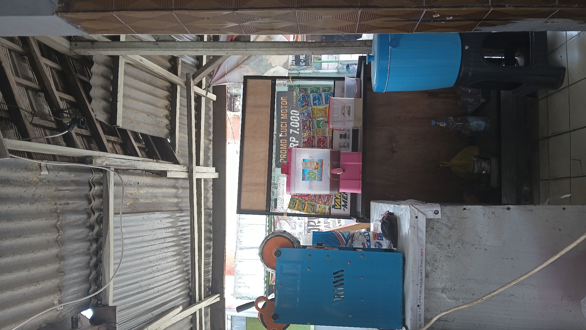This is a street-level, medium-shot photograph of a small roadside kiosk or warung, likely situated in the city of Dayeuhkolot, Indonesia. The kiosk appears to be set up outdoors under a corrugated metal roof supported by rough wooden beams. The overall impression is one of a functional, possibly temporary, vending stall.
In the foreground, to the left, a bright blue plastic sealing machine is prominent. It has orange circular elements attached to its top. To the right of this machine, on a light-colored surface that appears to be a covered counter or table, are several items. A dark fabric item, possibly a cloth or towel, rests on this surface. Behind this, a large, clear plastic beverage dispenser with a pink base holds a dark liquid, possibly tea or coffee. There are also other transparent plastic containers visible, one with a pale yellow liquid and another with a reddish liquid.
Further back, behind the counter, a wooden board serves as a backdrop. A sign is attached to this board, featuring Indonesian text that includes "PROMO CUCI MOTOR" (Motor Wash Promo) and "RP 7.000". Various packets and products are displayed behind the beverage dispenser and on shelves.
To the far right of the image, a tall, bright blue plastic water cooler with a lid sits on a black plastic stool. The wall to the right of the kiosk is tiled with a repetitive brown pattern.
The environment suggests an active commercial area, typical of roadside establishments in Indonesian cities. The lighting indicates it is daytime. There are no people visible in the frame. The flooring appears to be tiled. The overall condition of the kiosk and its surroundings suggests a utilitarian rather than a highly maintained setup, consistent with a local business.