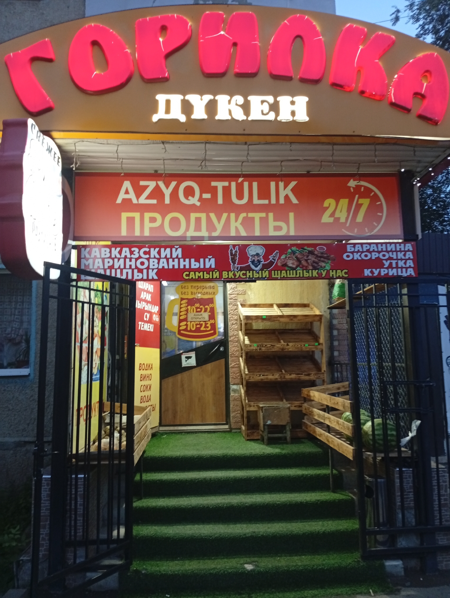 The image shows a storefront with a prominent sign displaying the Cyrillic word "Горнока" in large, red, illuminated letters. Below this, in white letters, is "ДУКЕН", indicating a market or shop. Further down, a red banner reads "AZYQ-TÚLIK" and "ПРОДУКТЫ", meaning "Food Products" in Kazakh and Russian respectively, with a "24/7" symbol indicating it is open around the clock.
The storefront itself has a dark brown facade with a glass door. To the left of the entrance, a black metal gate allows a glimpse of a shelf with various bottles, suggesting it might be a liquor store or a place selling beverages. To the right of the entrance, wooden shelves are set up, with some empty, indicating they are for display or sale. A section of the entrance has artificial turf covering the steps and a small area in front, giving it a green, grassy appearance.
On the red banner, there is an illustration of a cartoonish man in traditional attire holding a kebab skewer. Below this illustration, text in Russian says "ВКУСНЫЙ ШАШЛЫК У НАС", meaning "Delicious Shashlik (kebab) at our place." To the right of this, another banner lists various meats: "БАРАНИНА" (mutton), "ОКОРОЧКА" (ham/drumstick), "УТКА" (duck), and "КУРИЦА" (chicken).
On the left side of the entrance, a vertical yellow sign lists various drinks: "ВОДКА" (vodka), "ВИНО" (wine), "СОКИ" (juices), and "ВОДА" (water).
The overall impression is a local food and beverage shop, possibly a deli or a small grocery store, in a city. The presence of Kazakh and Russian languages on the signs suggests it is located in a region where both languages are commonly used, such as Oral, Kazakhstan.