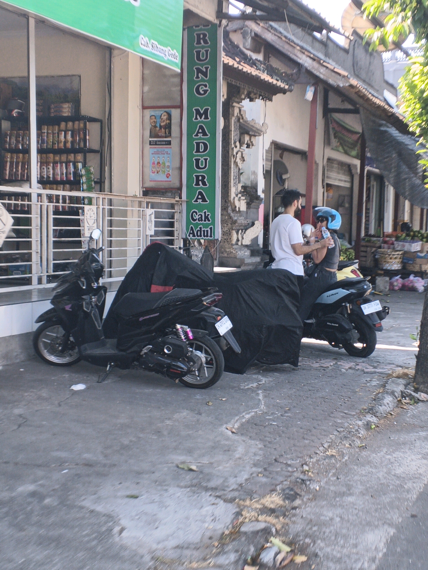 In front of a shop with a green awning and the text "Cak Sitibang Gede" visible, two motorcycles are parked on a concrete sidewalk. The motorcycle in the foreground is black and partially covered by a black tarp. The motorcycle behind it is also black and is mostly covered by the same black tarp, revealing its rear wheel and license plate. A man in a white t-shirt and dark pants stands to the right of the motorcycles, facing a woman who is sitting on the rear motorcycle, wearing a dark top and dark pants, and a blue helmet. She is holding a bottle of water and appears to be drinking from it, with the man holding the bottle. The shop facade has shelves stocked with goods and posters on the wall. A vertical green sign with white text reading "RUNG MADURA Cak Adul" is also visible on the right side of the shop entrance. The environment suggests a street in Abiansemal, Indonesia, with typical urban elements and vegetation.