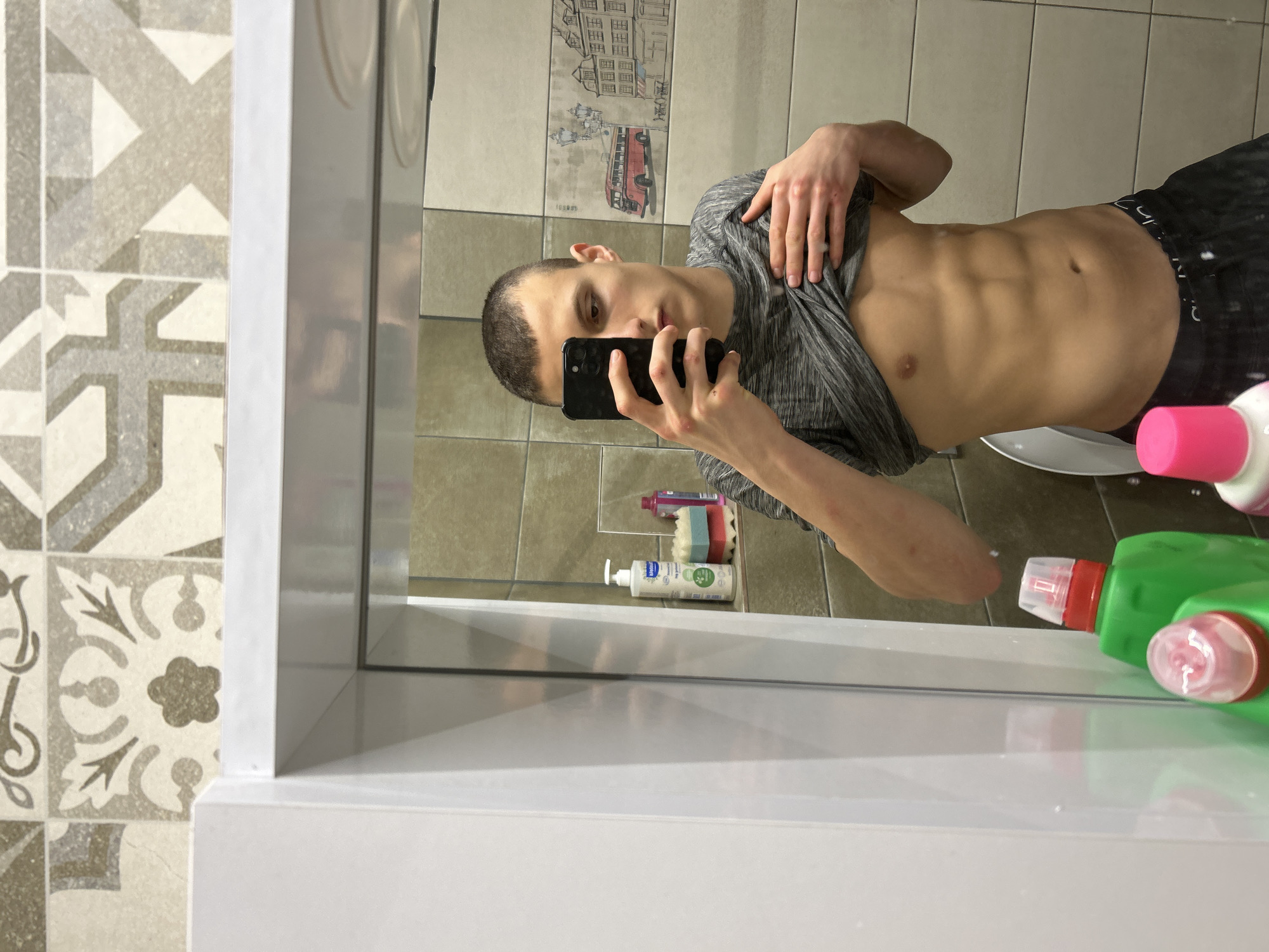 A young man takes a mirror selfie in a bathroom. He is fair-skinned with short, dark hair. He is wearing a gray t-shirt that he is pulling up to expose his abdomen, revealing defined abdominal muscles. He is also wearing dark shorts. He is holding a black smartphone in his right hand. The background shows bathroom tiles, some with decorative patterns. Various bottles of cleaning products are visible on the counter near the bottom right of the image. The photograph appears to be taken in a residential setting. It is not possible to determine if the location is in Burgas, Bulgaria based solely on the image.