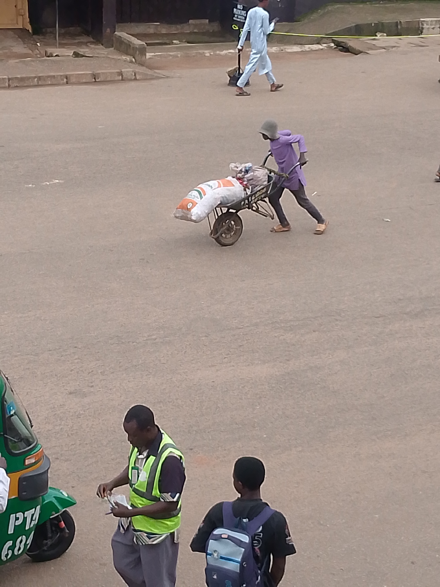 In Bukuru, Nigeria, a man in a purple shirt and dark pants pushes a wheelbarrow laden with two large white sacks. He wears a gray hat. In the background, another man in a light blue garment walks away from the camera. Closer to the foreground, a man wearing a bright green safety vest over a dark shirt and gray pants stands next to a green auto-rickshaw and appears to be holding a document and a pen. To the right, a young person wearing a backpack faces away from the camera. The scene takes place on a paved surface, likely a street or parking lot, with some debris visible.